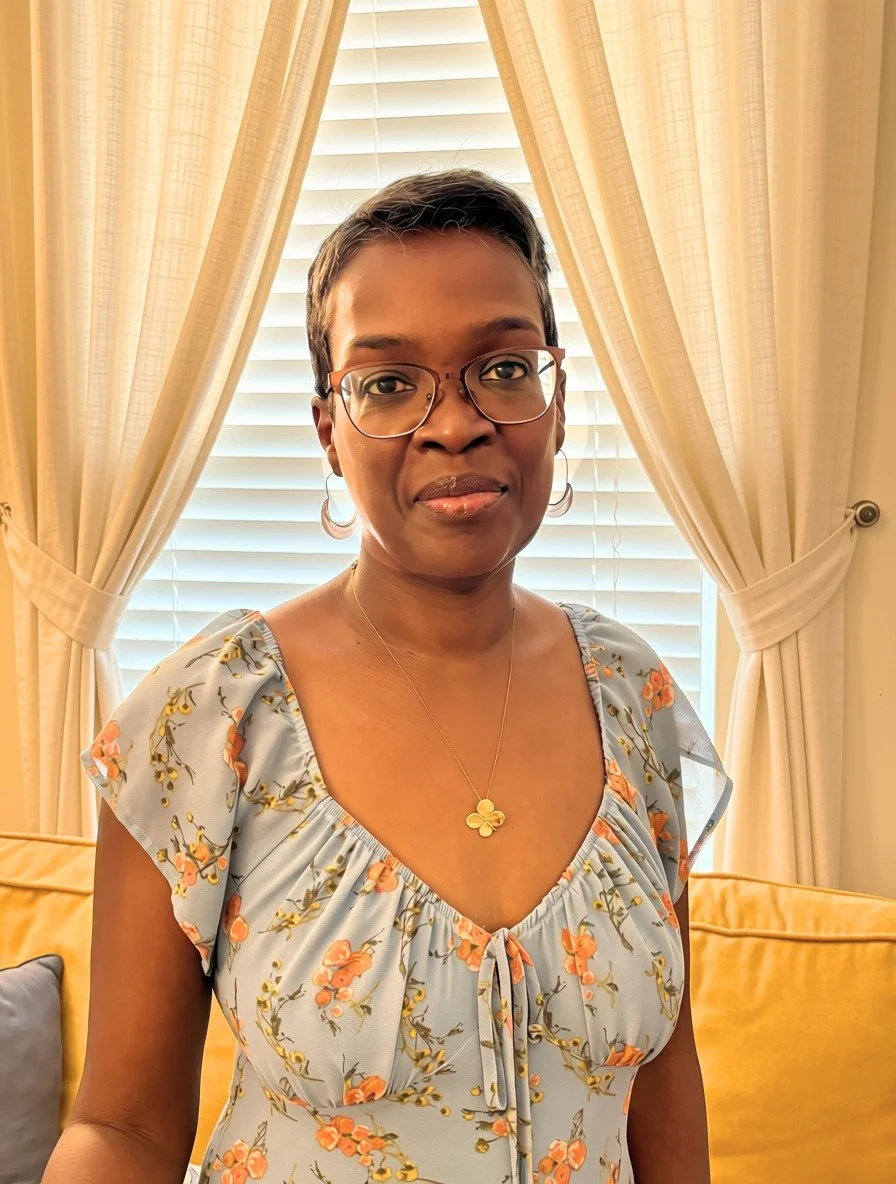A woman with short dark hair, glasses, and hoop earrings standing in front of cream-colored curtains and blinds, wearing a floral dress with a tie at the front and a gold four-leaf clover necklace.