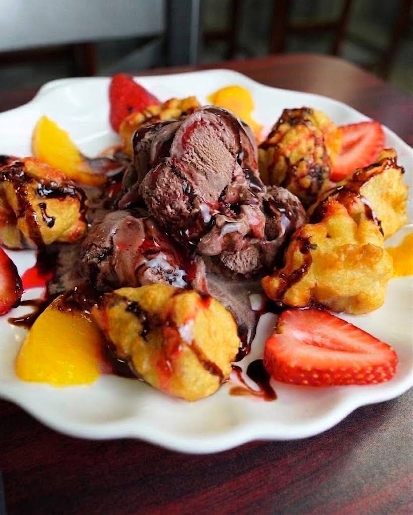 A bowl of fried plantains topped with chocolate ice cream, strawberry slices, and fruit slices, garnished with chocolate syrup.