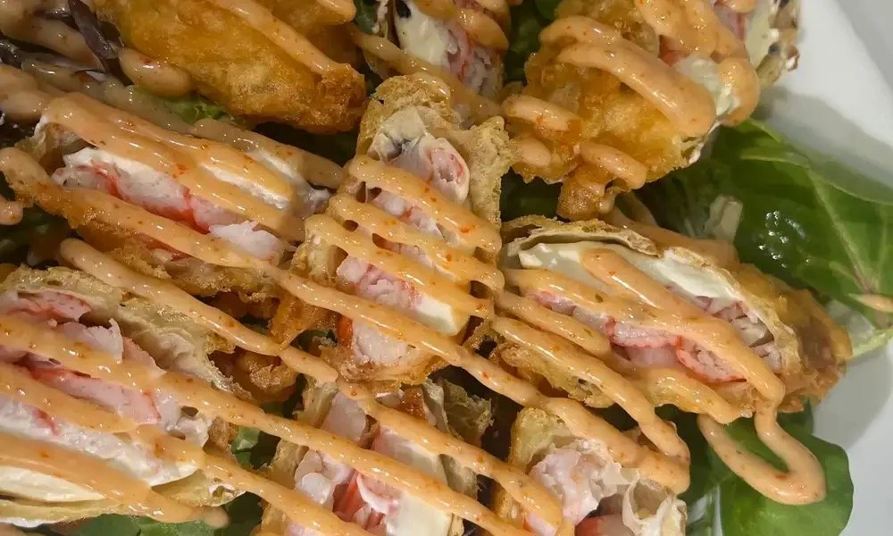 Close-up of sushi rolls topped with orange sauce, crispy fried exterior, and a drizzle of additional sauce on a bed of green leafy garnishes.