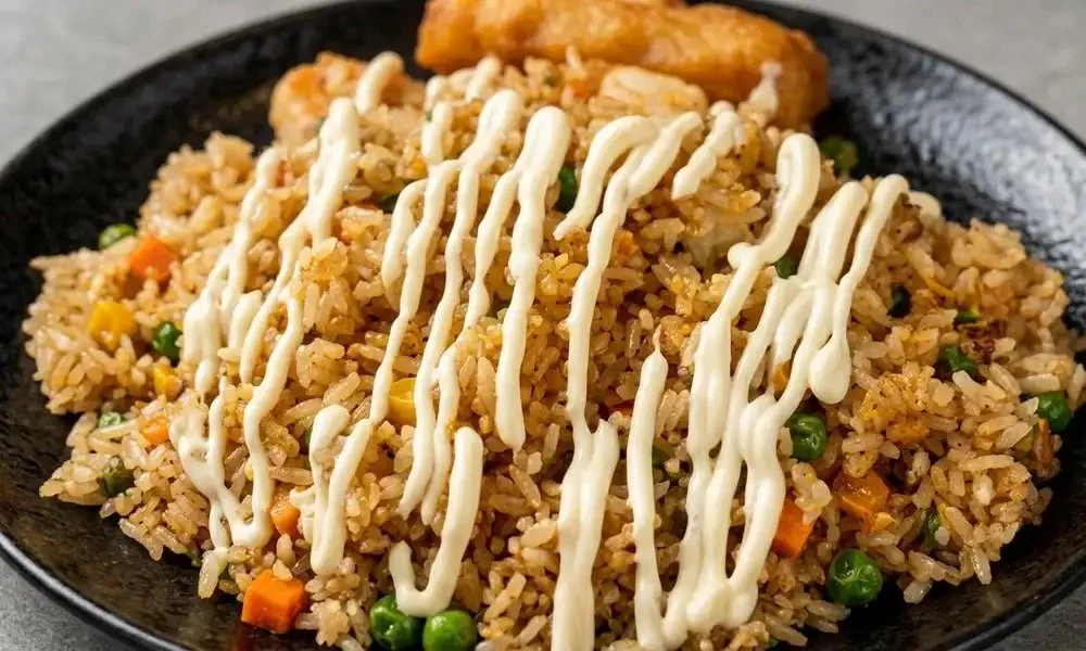 Fried rice topped with mayonnaise in a zigzag pattern, served with a slice of fried plantain on a black plate.