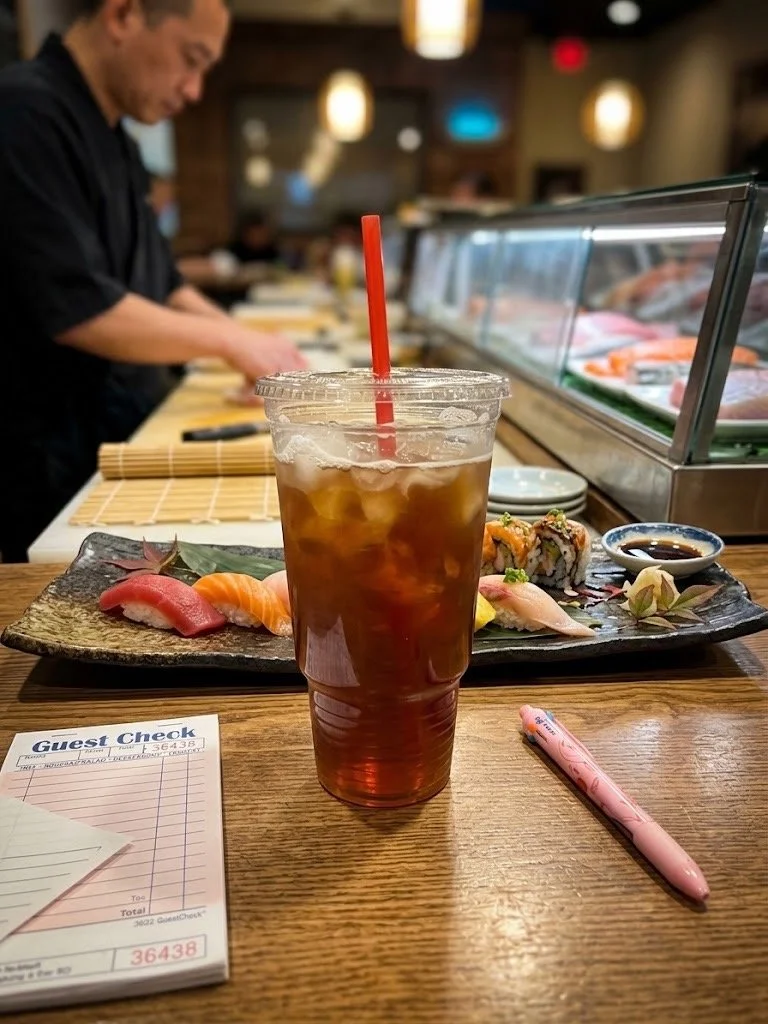 A plastic cup of iced tea with a red straw on a wooden counter at a sushi restaurant. Behind, a sushi chef prepares on a counter with sushi and a guest check receipt on the table.