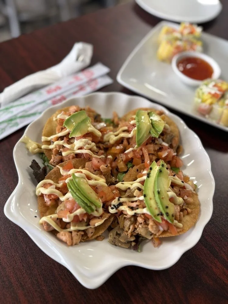 Plate of chicken tacos topped with sliced avocado, chopped tomatoes, mayonnaise, and black sesame seeds, with side of spring rolls and dipping sauce.