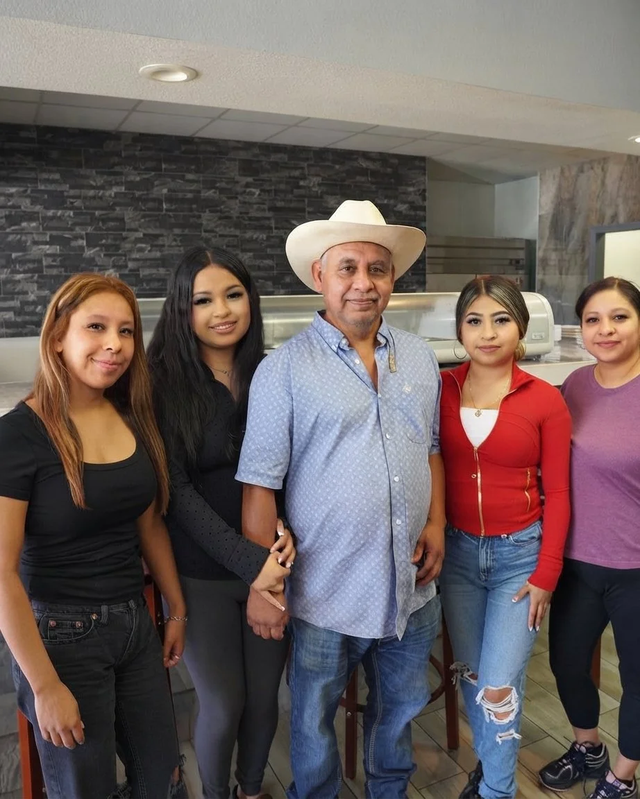Group of five people standing together in a restaurant, with the man in the middle wearing a cowboy hat and a light blue shirt.