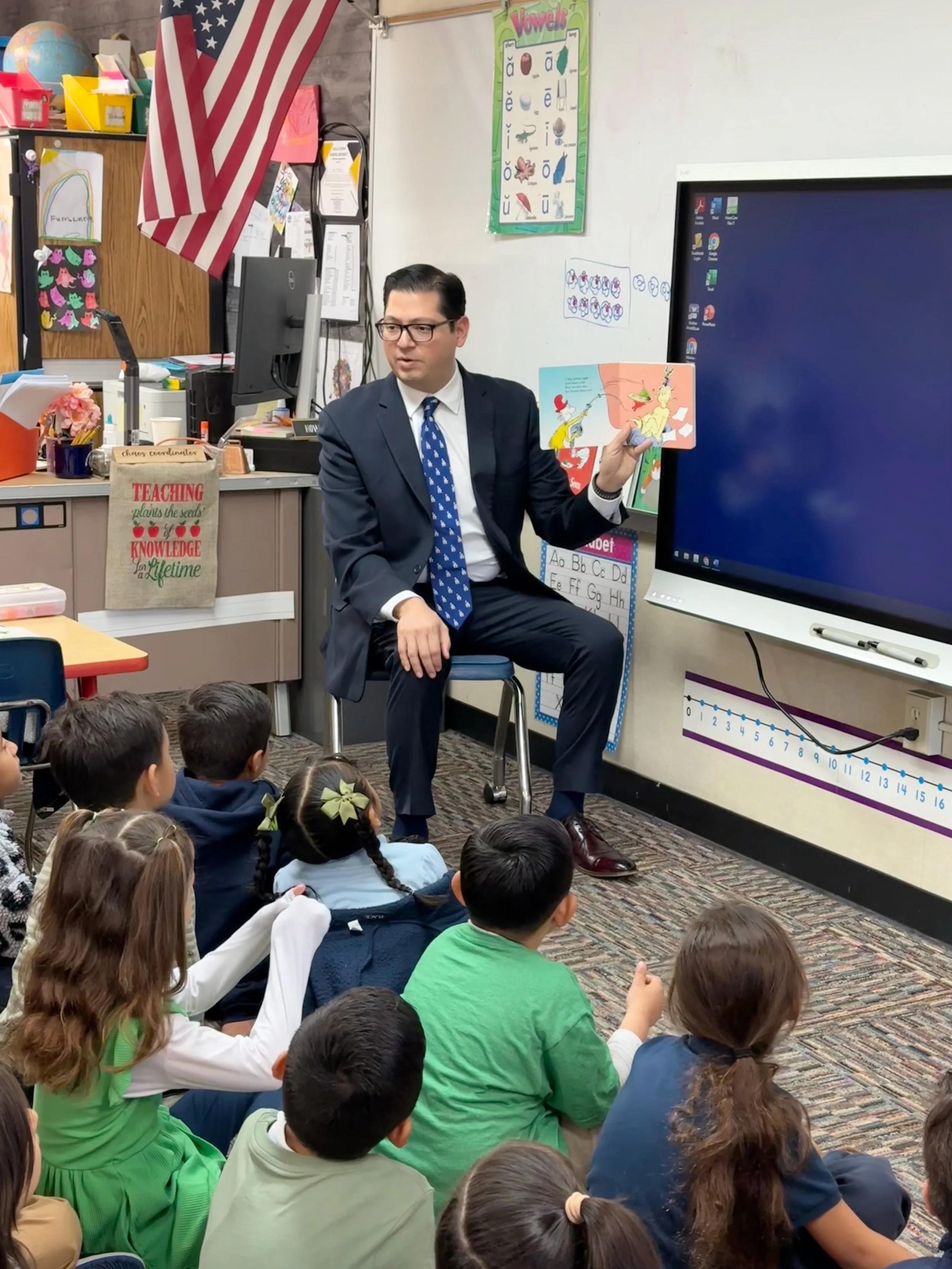 A man in a suit, sitting in a classroom reading a book to children.