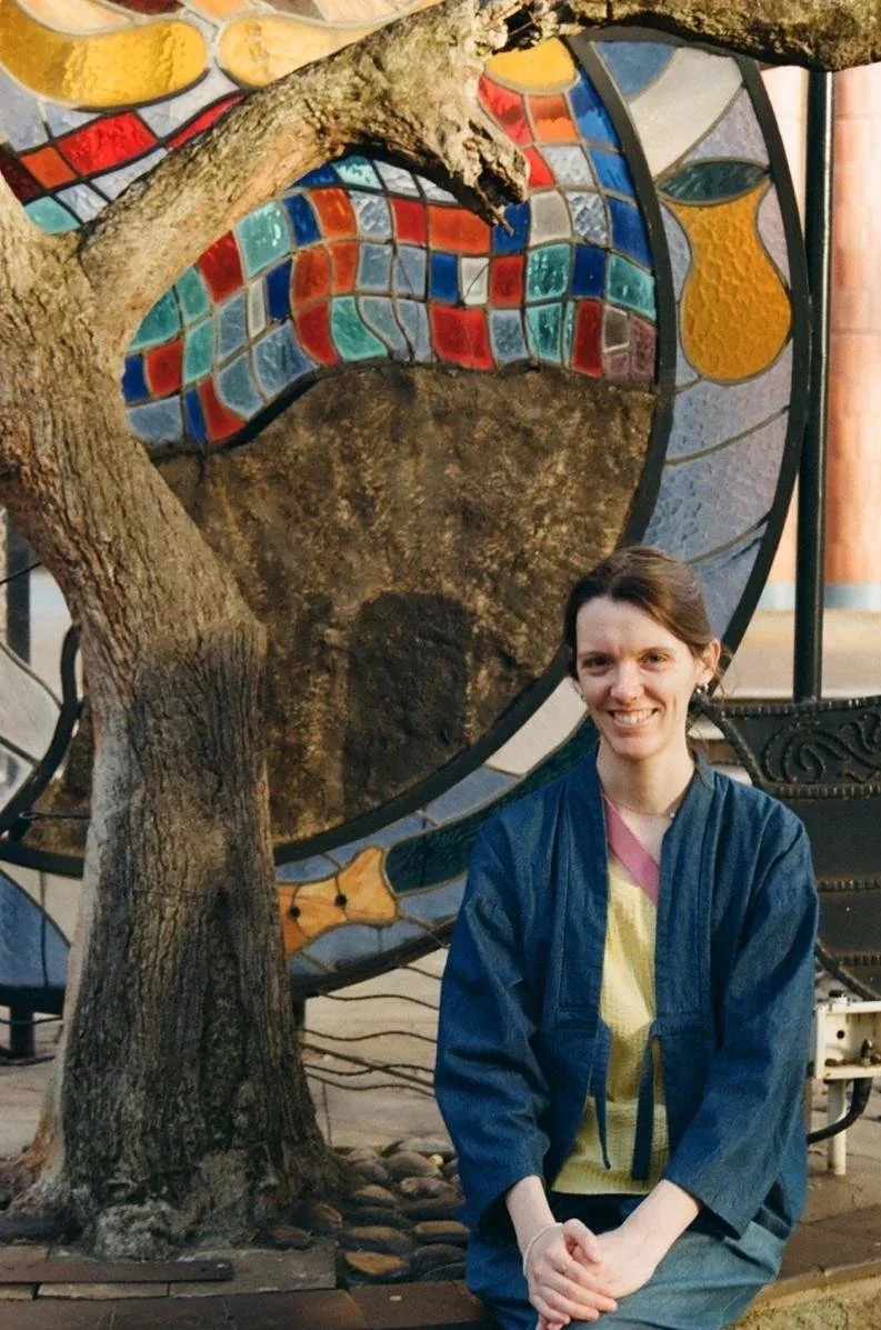 Holly wears a denim jacket with a yellow top. She sits in front of a tree and a sculpture with coloured glass.