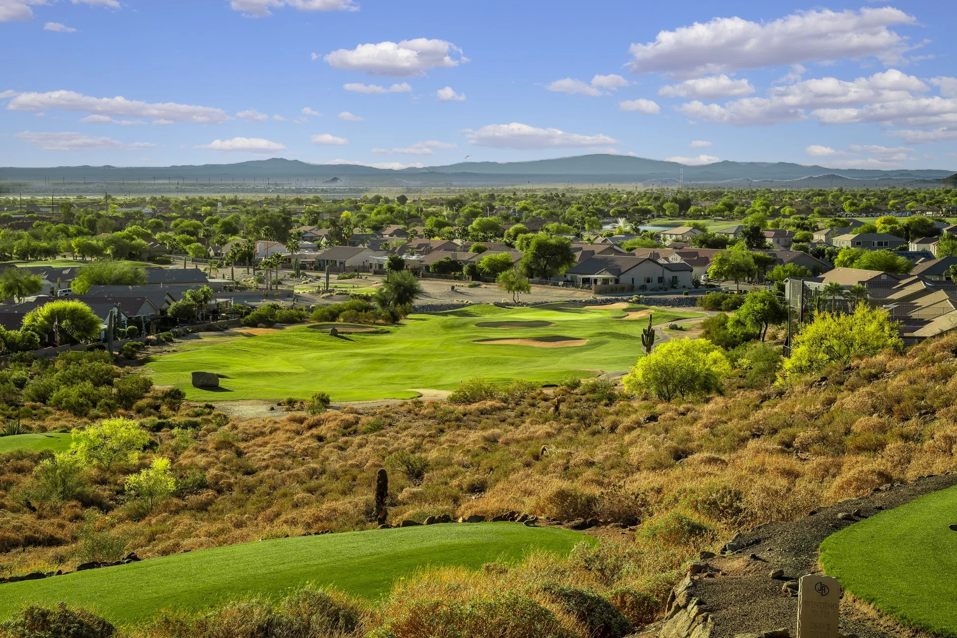 Landscape view of a golf course with neatly maintained green, surrounded by desert plants and rocky terrain. Residential houses are visible in the background under a bright blue sky with scattered white clouds, and mountains can be seen in the distance.