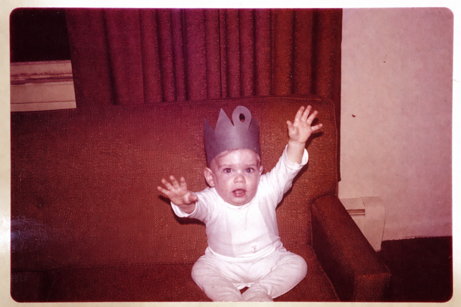 A young child sitting on a red couch, wearing a paper crown, with arms raised and a surprised or confused expression.