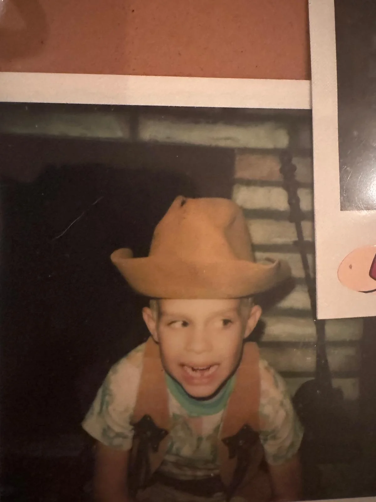A young boy wearing a cowboy hat and a backpack, smiling and looking to the side, in front of a staircase.