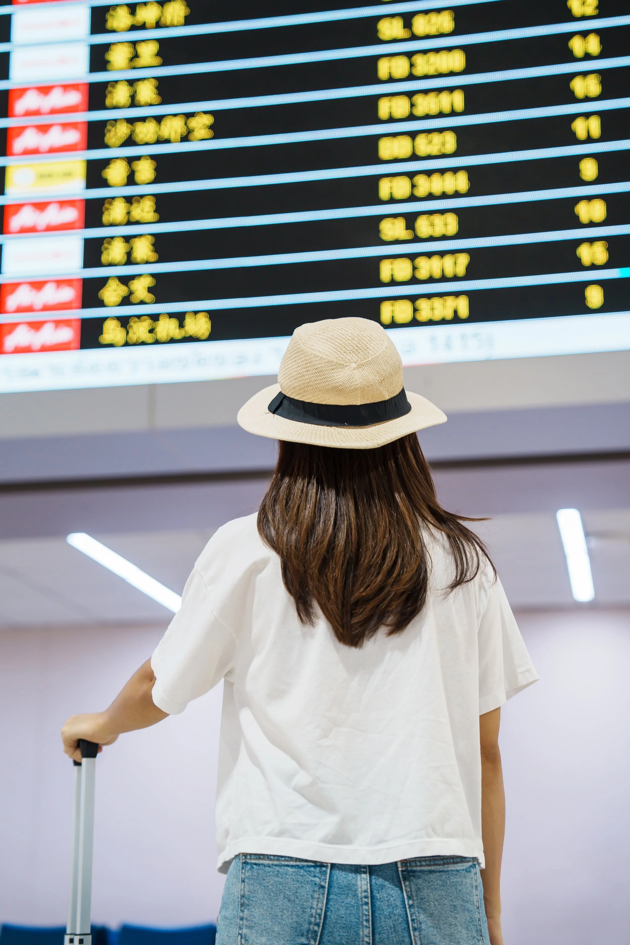 Person wearing a straw hat with black ribbon, a white shirt, and jeans, walking in an airport terminal with an electronic departure board in the background.