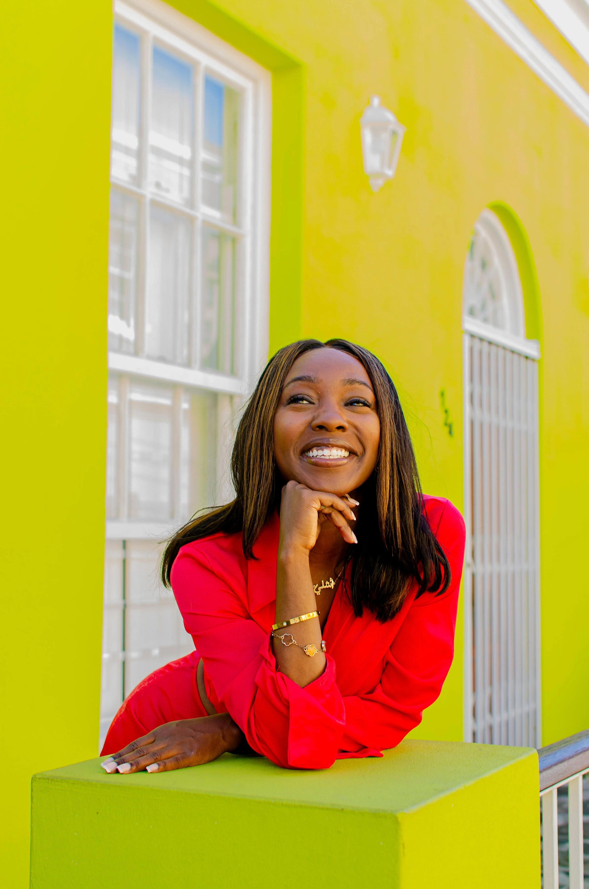 A woman with long dark hair in a red blazer, smiling and resting her chin on her hand, sitting at a bright lime green table in a colorful room with yellow walls and white-trimmed windows.