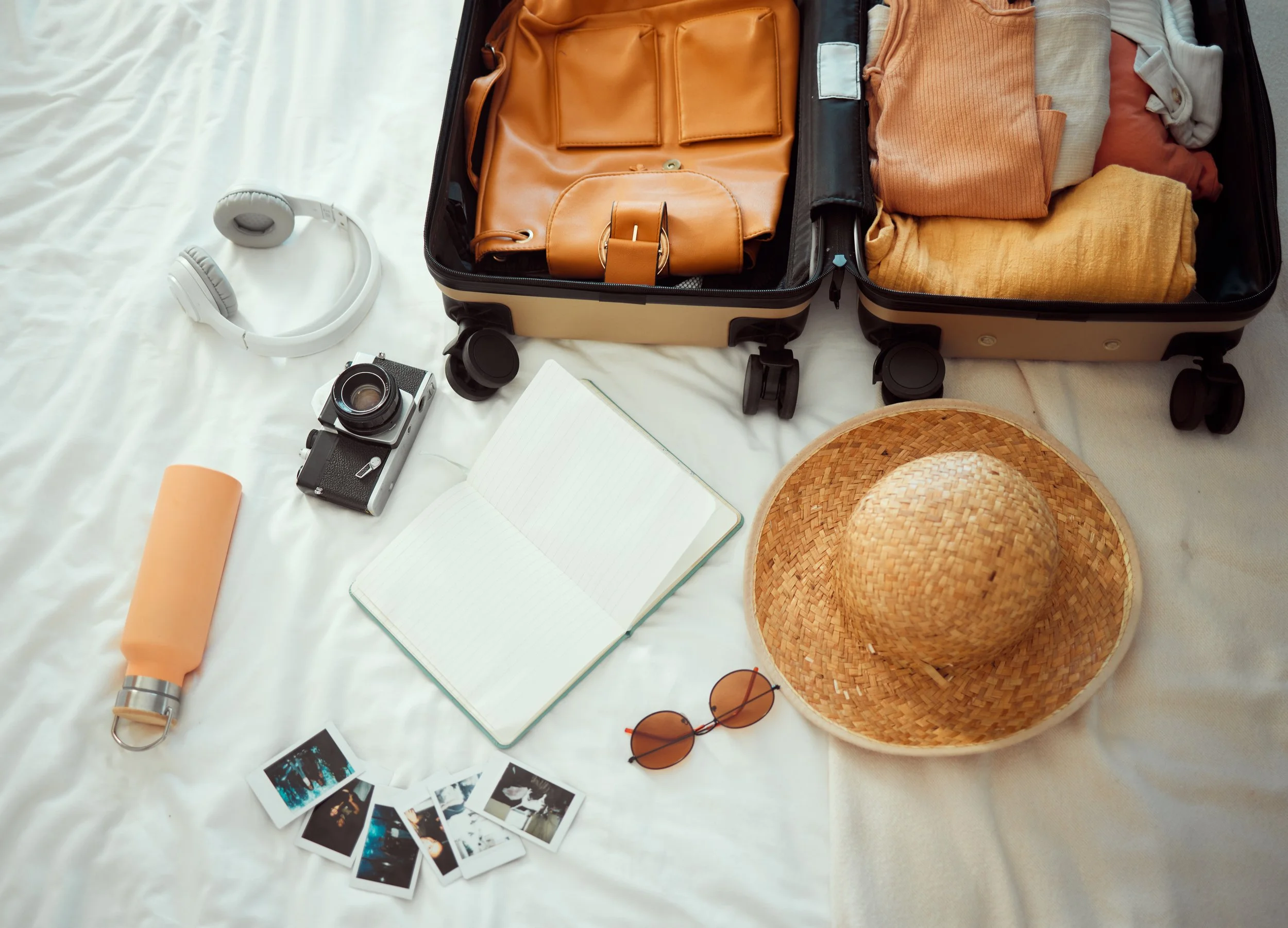 An open suitcase containing clothes, a brown leather handbag, a notebook, and a straw hat. Surrounding the suitcase on a white bed are white headphones, a vintage camera, an orange water bottle, sunglasses, and some printed photographs.