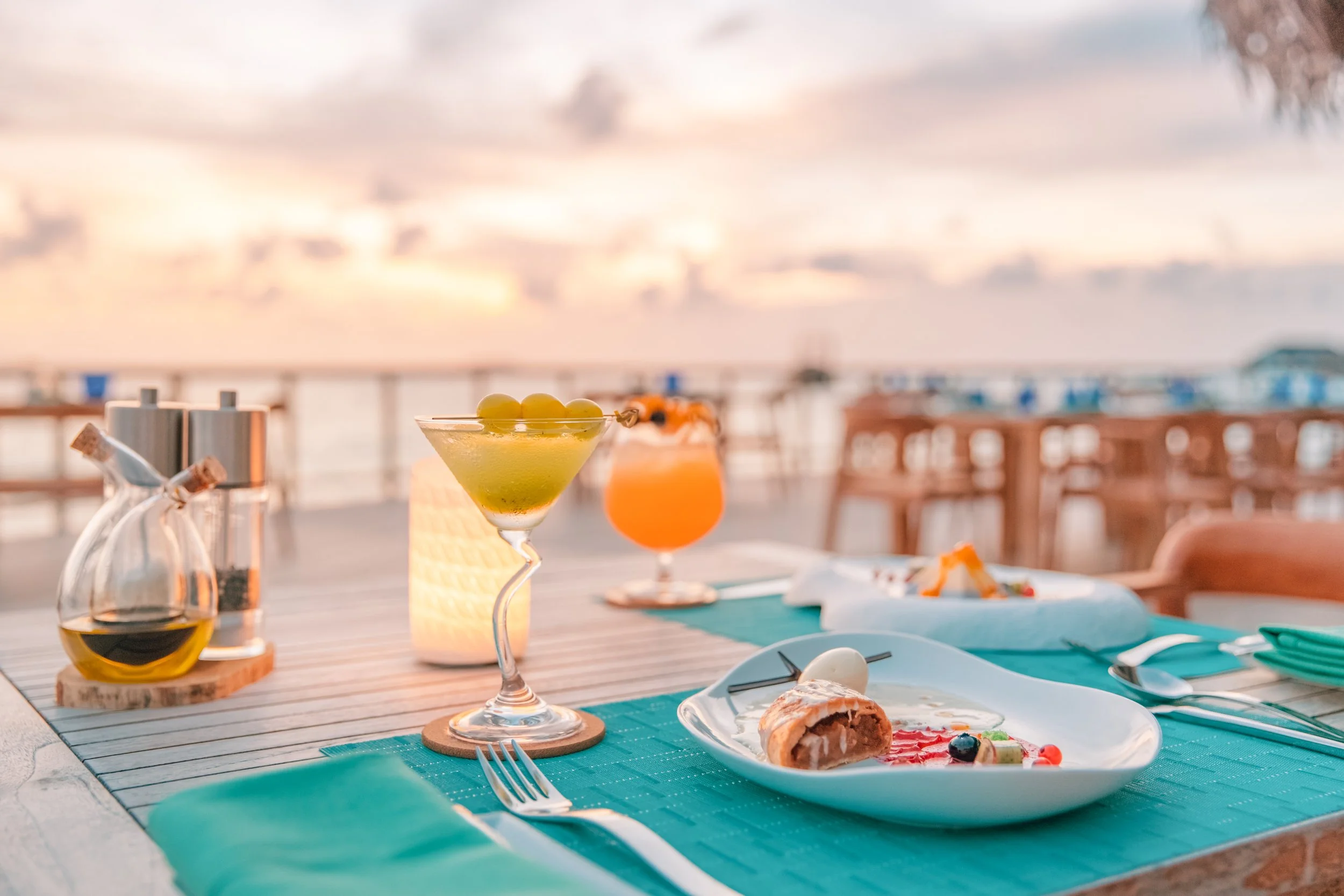 Table set with drinks and food on an outdoor patio during sunset, featuring a cocktail with green olives, a glass of orange beverage, plates with appetizers, and a rolled pastry.