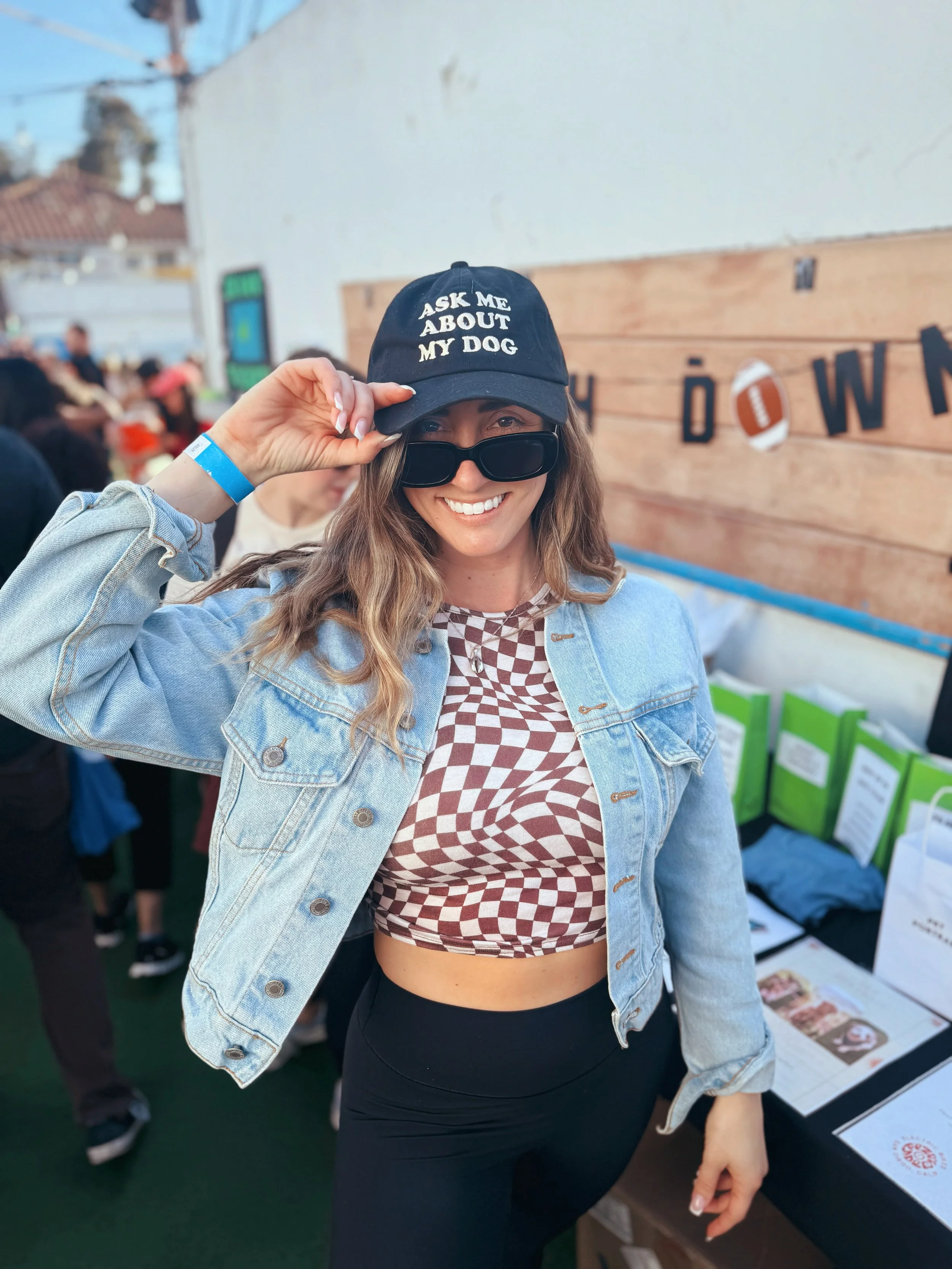 A smiling woman wearing black sunglasses and a denim jacket, holding a black baseball cap that reads 'Ask Me About My Dog,' standing in front of an outdoor booth.
