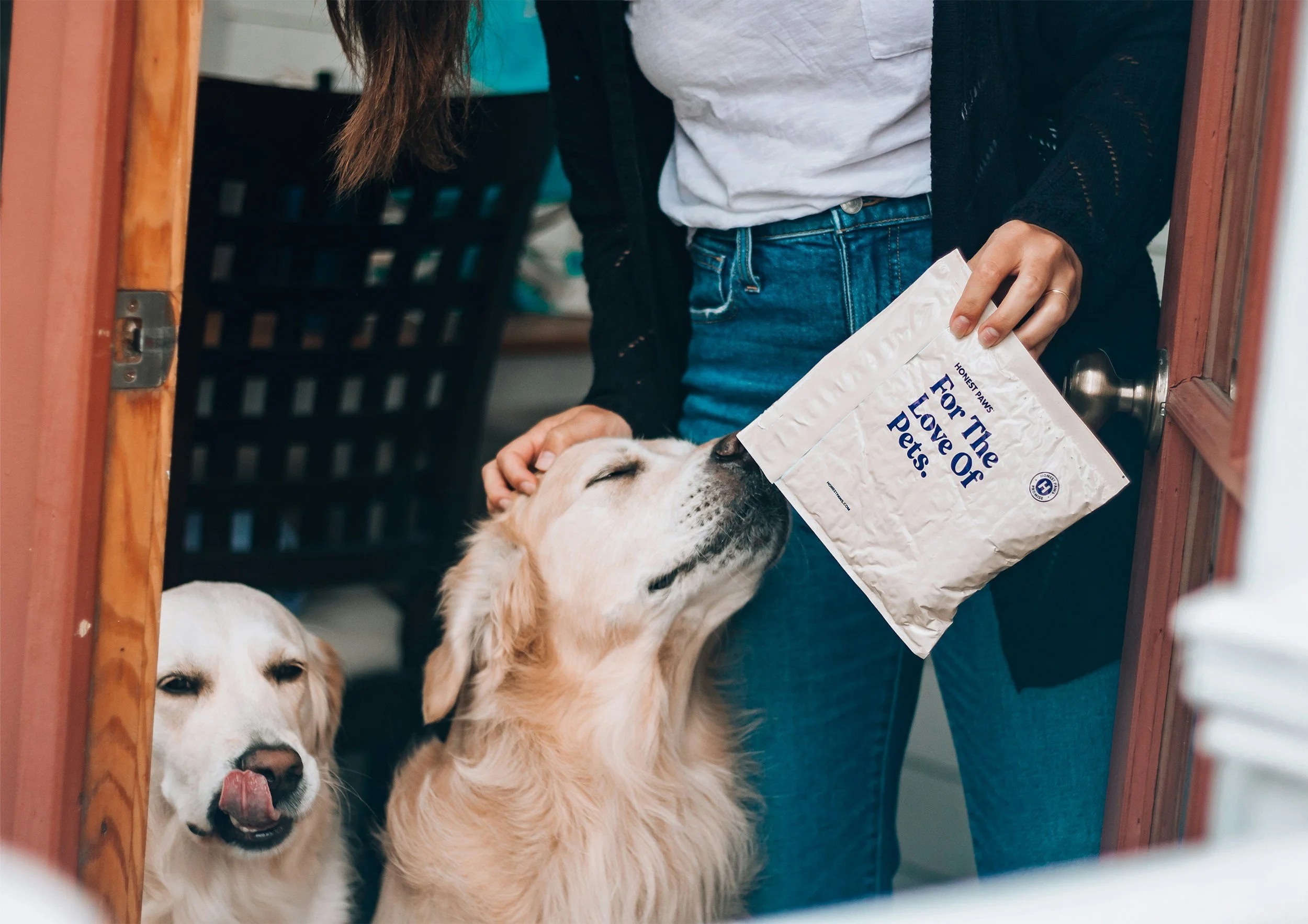 Two dogs, one golden retriever with closed eyes and the other with a tongue out, are being fed treats from a person's hand through a doorway.