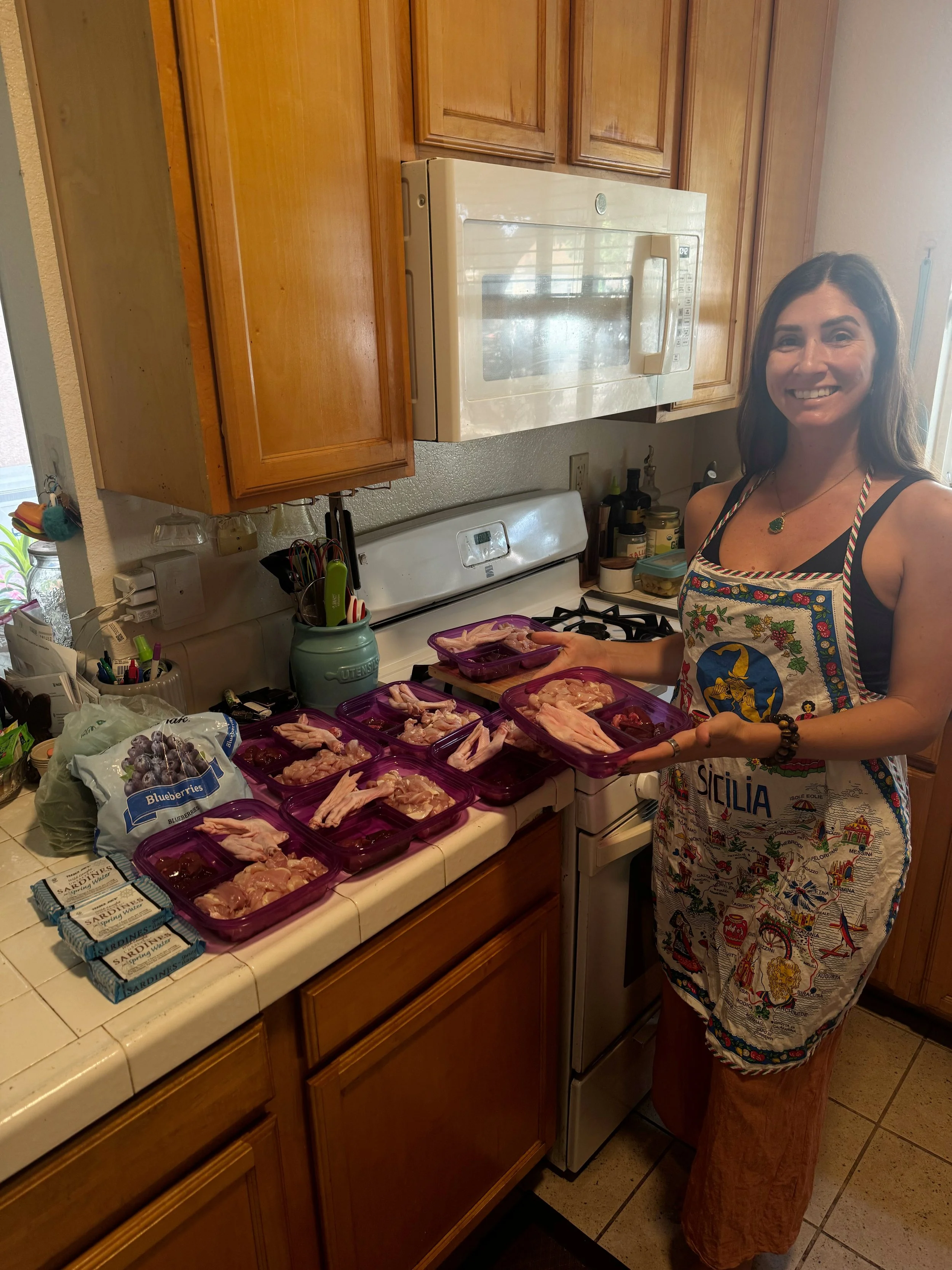 A woman wearing a colorful apron with various images on it, standing in a kitchen, holding containers of raw chicken on a countertop. The kitchen has wooden cabinets, a white microwave, and various kitchen utensils and ingredients visible.