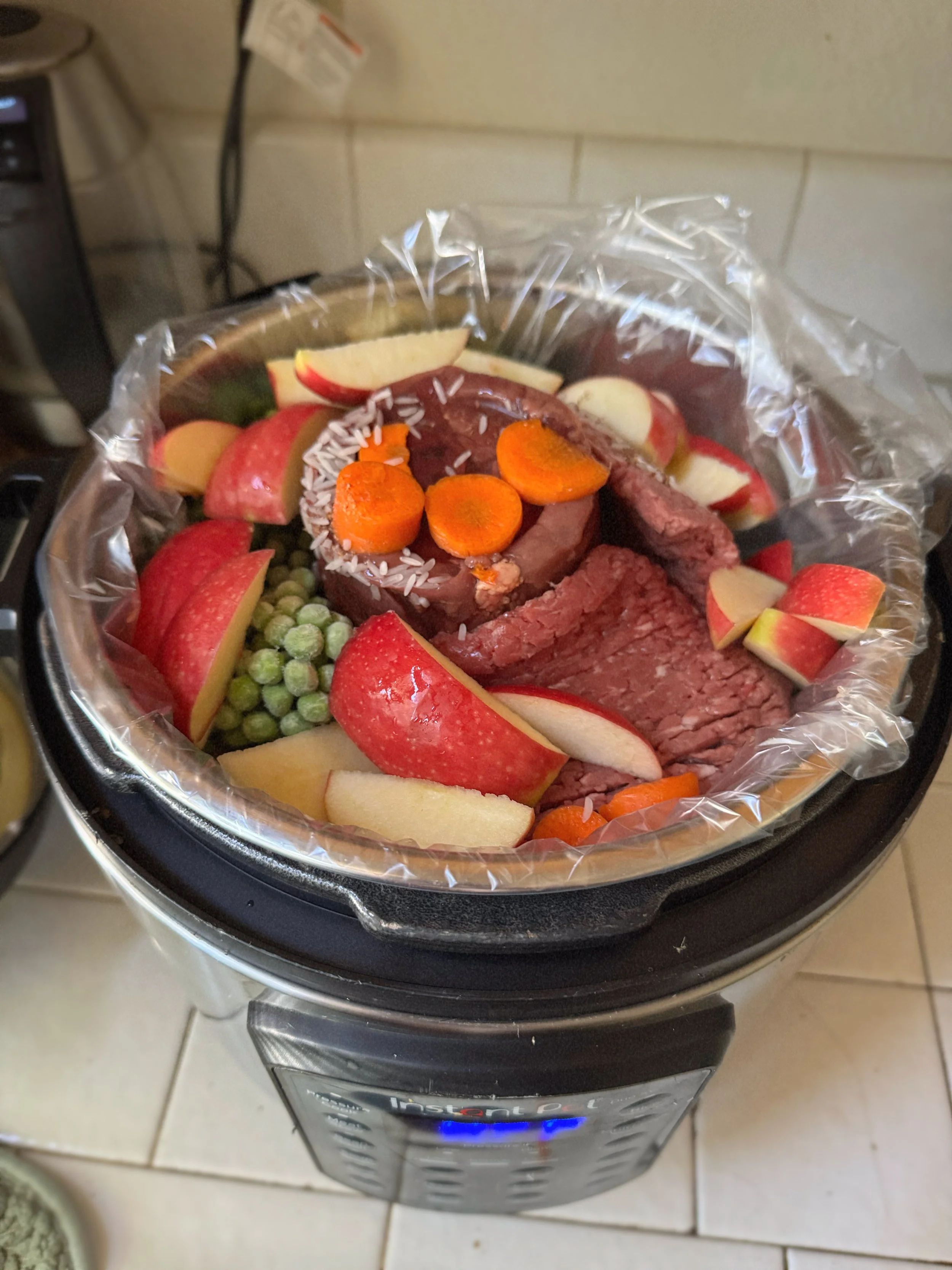 A black Instant Pot electric pressure cooker with sliced apples, carrots, green peas, and raw ground beef inside, covered with plastic wrap, on a kitchen countertop. A lightly cooked meal prep for the dogs.