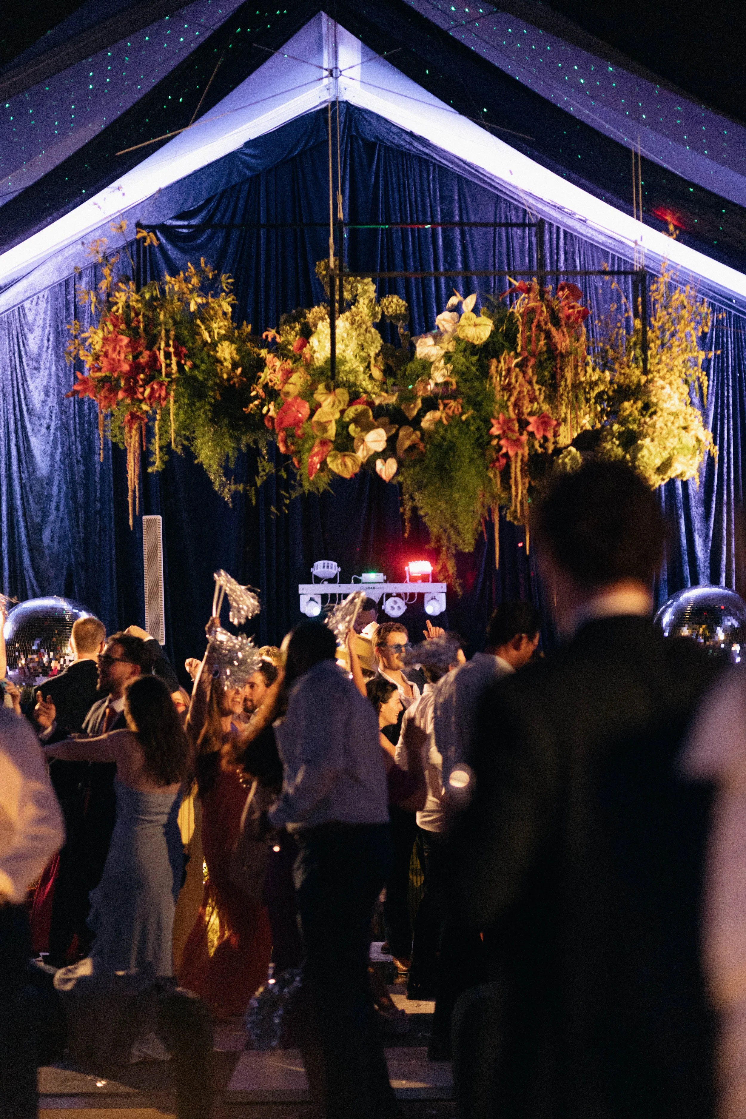 A party scene with people dancing under a decorated tent featuring hanging flowers and string lights.