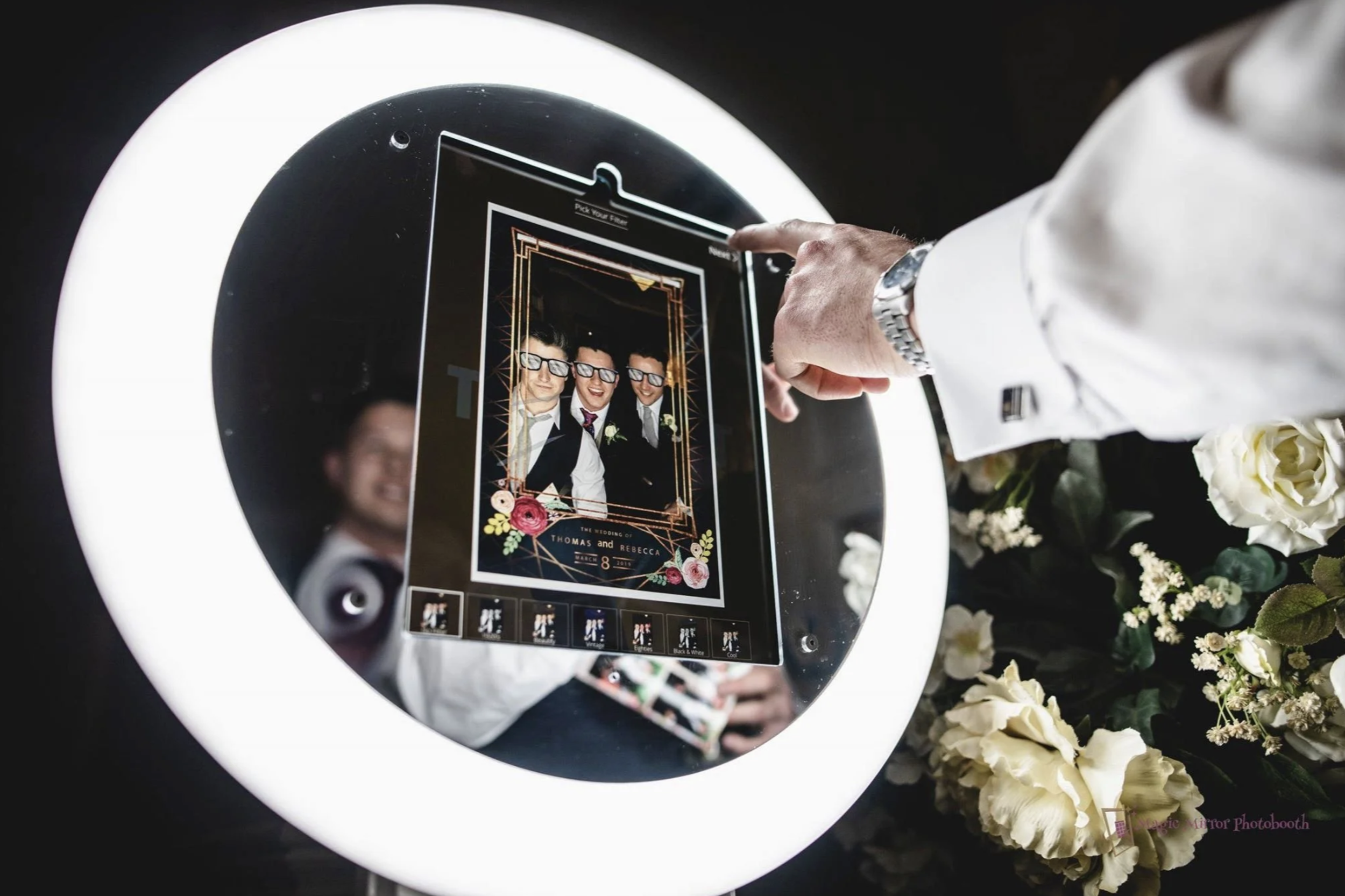 A person is using a touchscreen device into which a wedding invitation is displayed, surrounded by white flowers.
