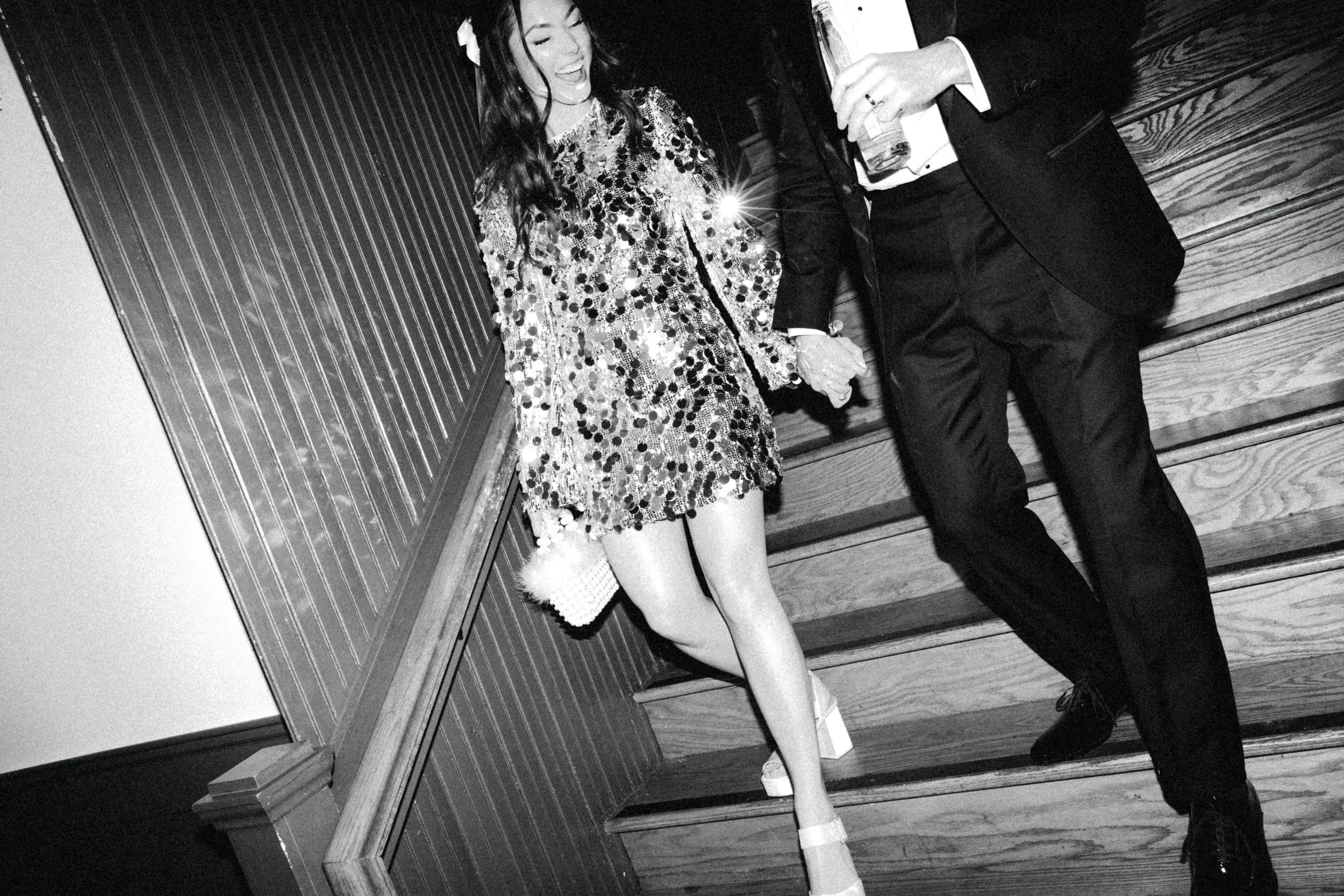 A woman in a shiny, sparkly dress sitting on a step, smiling and talking with a man in a suit, who is holding a drink. They are sitting on wooden stairs against a wooden wall.