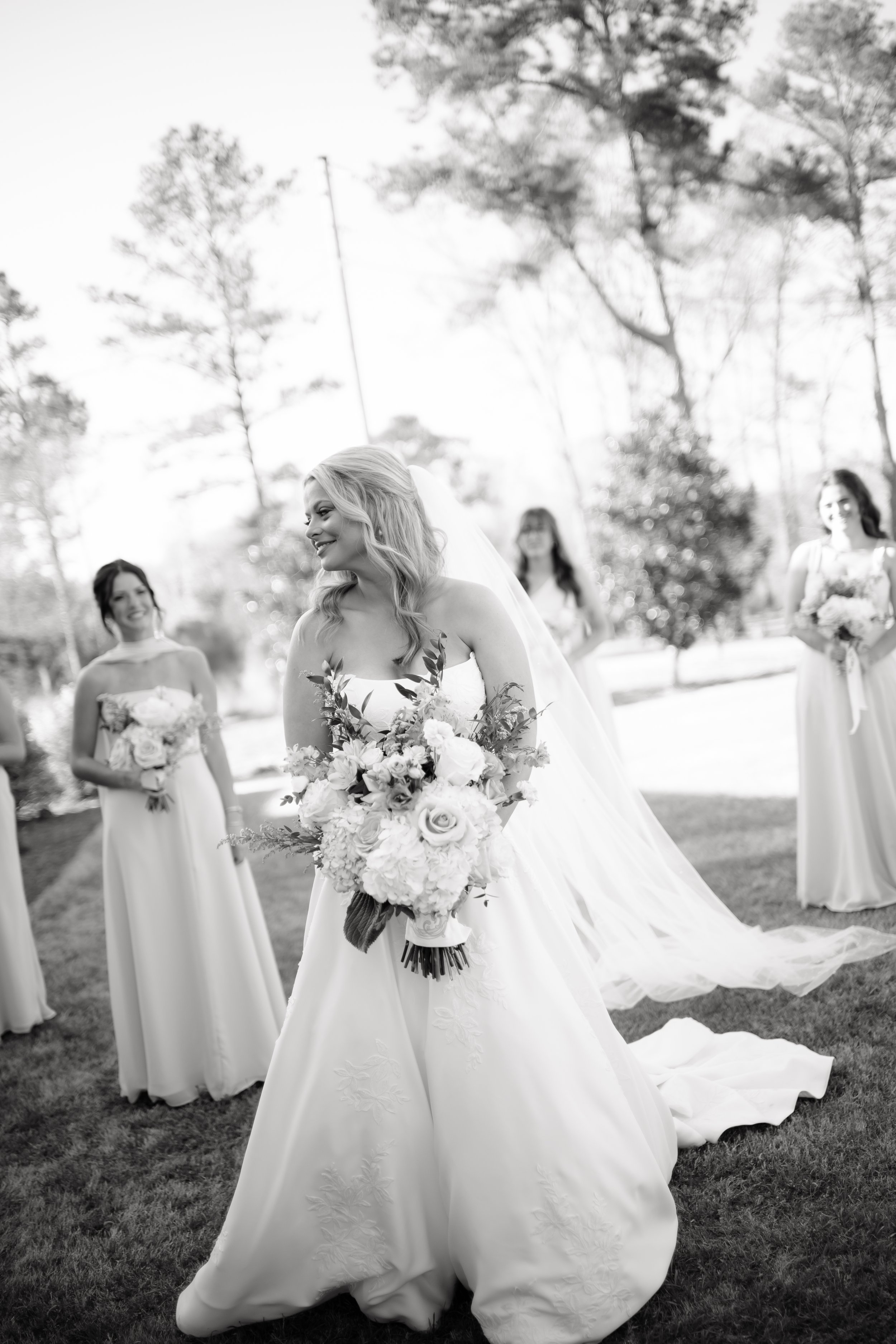 Black-and-white photo of a bride holding a bouquet of flowers, smiling, with three bridesmaids standing in the background outdoors on a grassy area with trees.