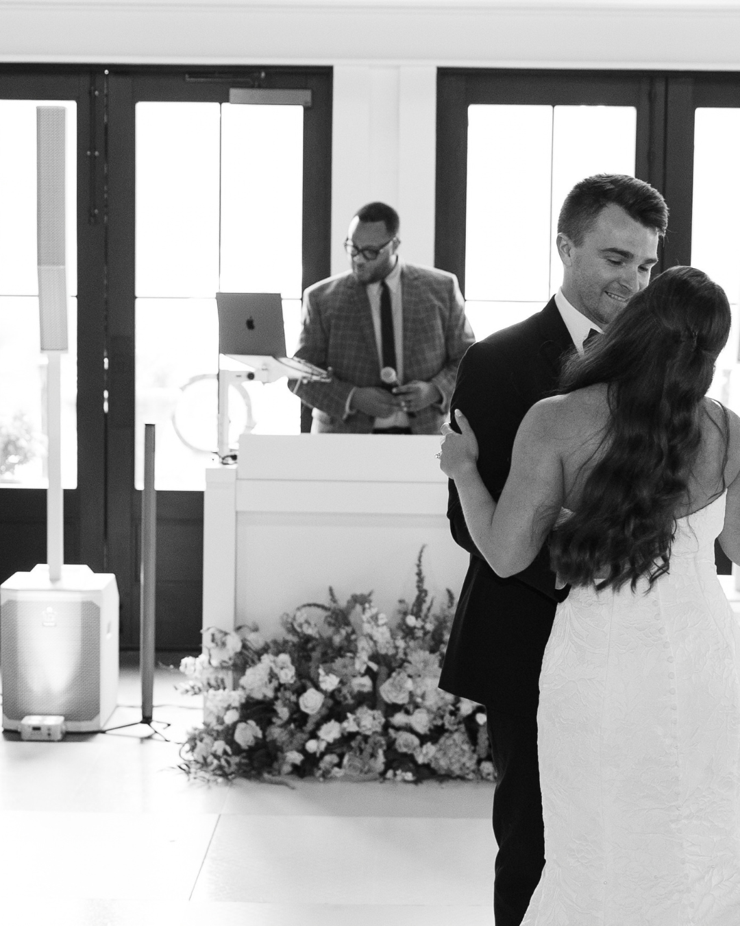 A black-and-white photo of a wedding reception, showing a bride and groom dancing together in the foreground with the groom smiling. In the background, a man in a checked suit, glasses, and tie, holding a microphone, stands near a DJ setup with a laptop, with large windows behind him letting in natural light.
