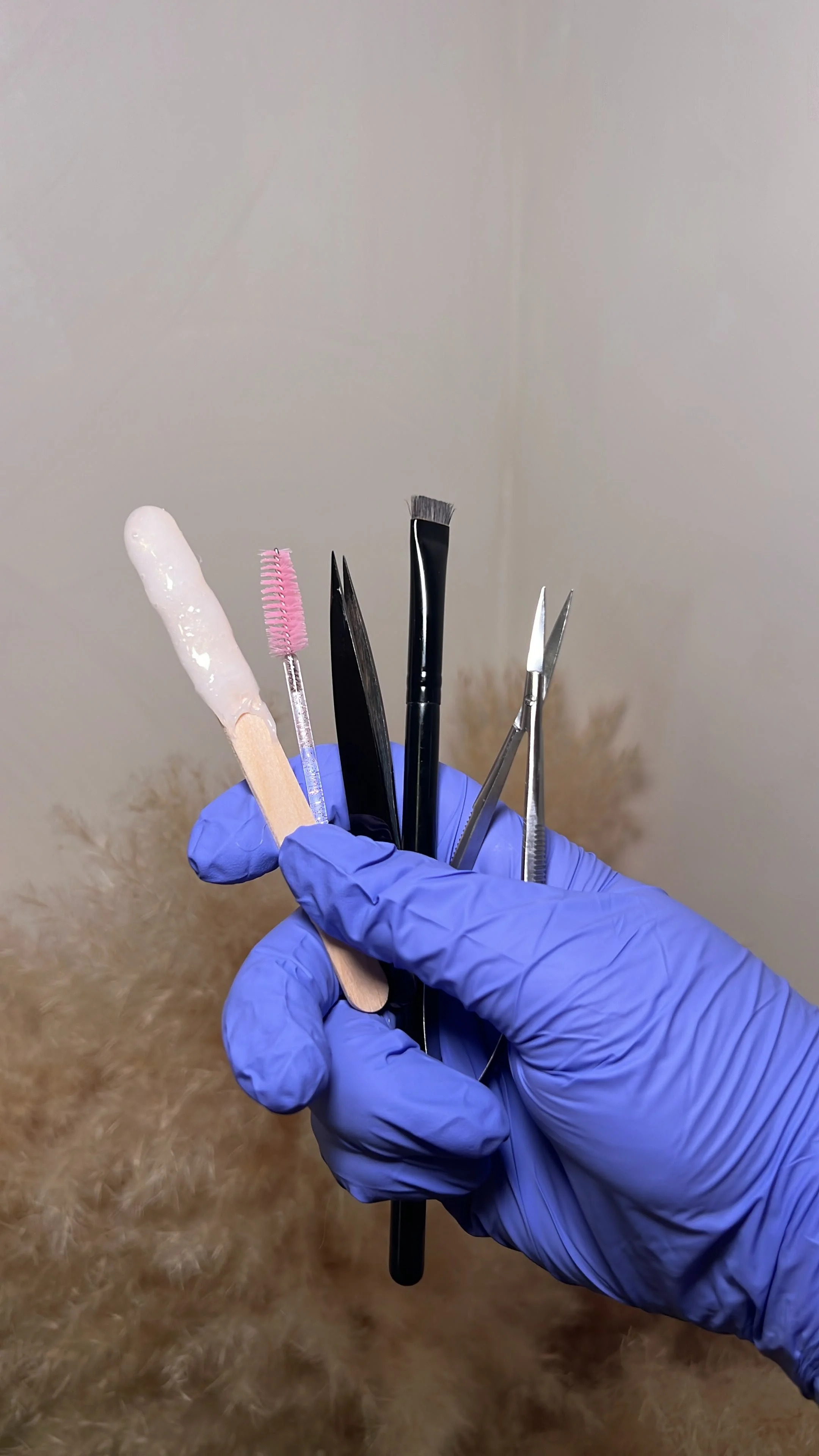 Person wearing a blue glove holding a collection of grooming tools, including a wooden stick with wax, a pink spoolie, black tweezers, a black brush, and small scissors.