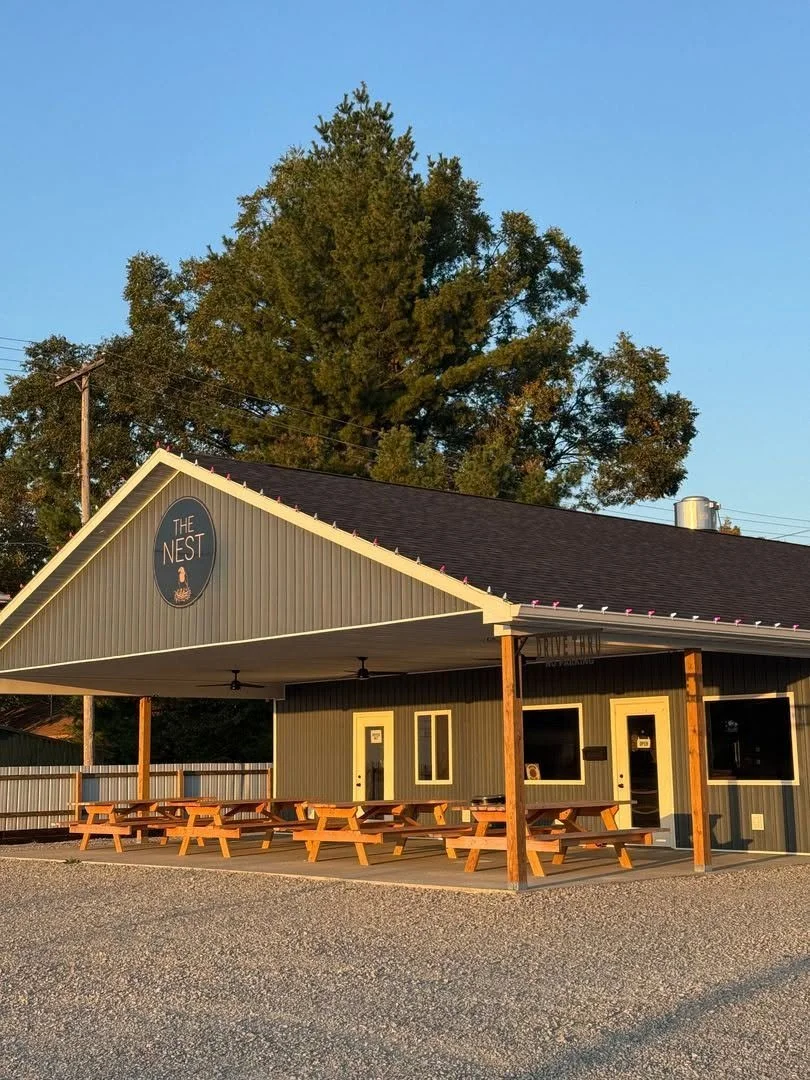 A small building named The Nest with outdoor picnic tables, surrounded by a gravel parking lot and trees, under a clear blue sky.
