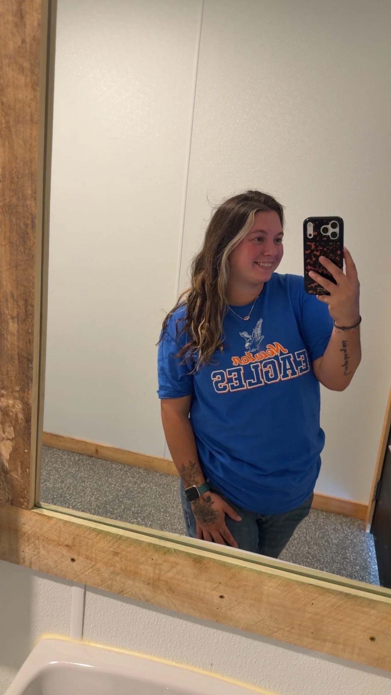 A young woman taking a selfie in a bathroom mirror. She is wearing a blue shirt with the words "New Orleans Falcons" on it, has long wavy hair, and tattoos on her arm.