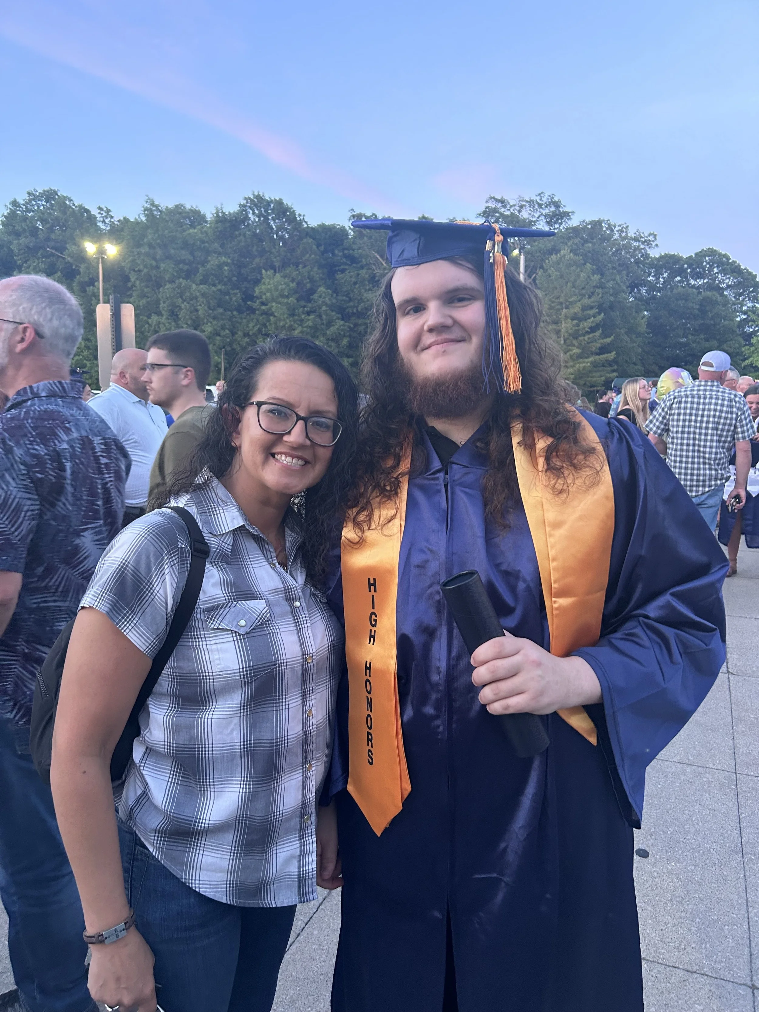 A young man wearing a graduation cap and gown with a gold stole, holding a diploma, standing next to a woman at an outdoor graduation ceremony with many people in the background.