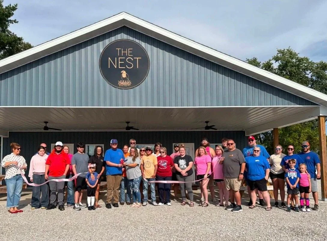Group of people at a ribbon-cutting event in front of a blue building with a sign that reads 'The NEST' and shows a nest with a bird on top.