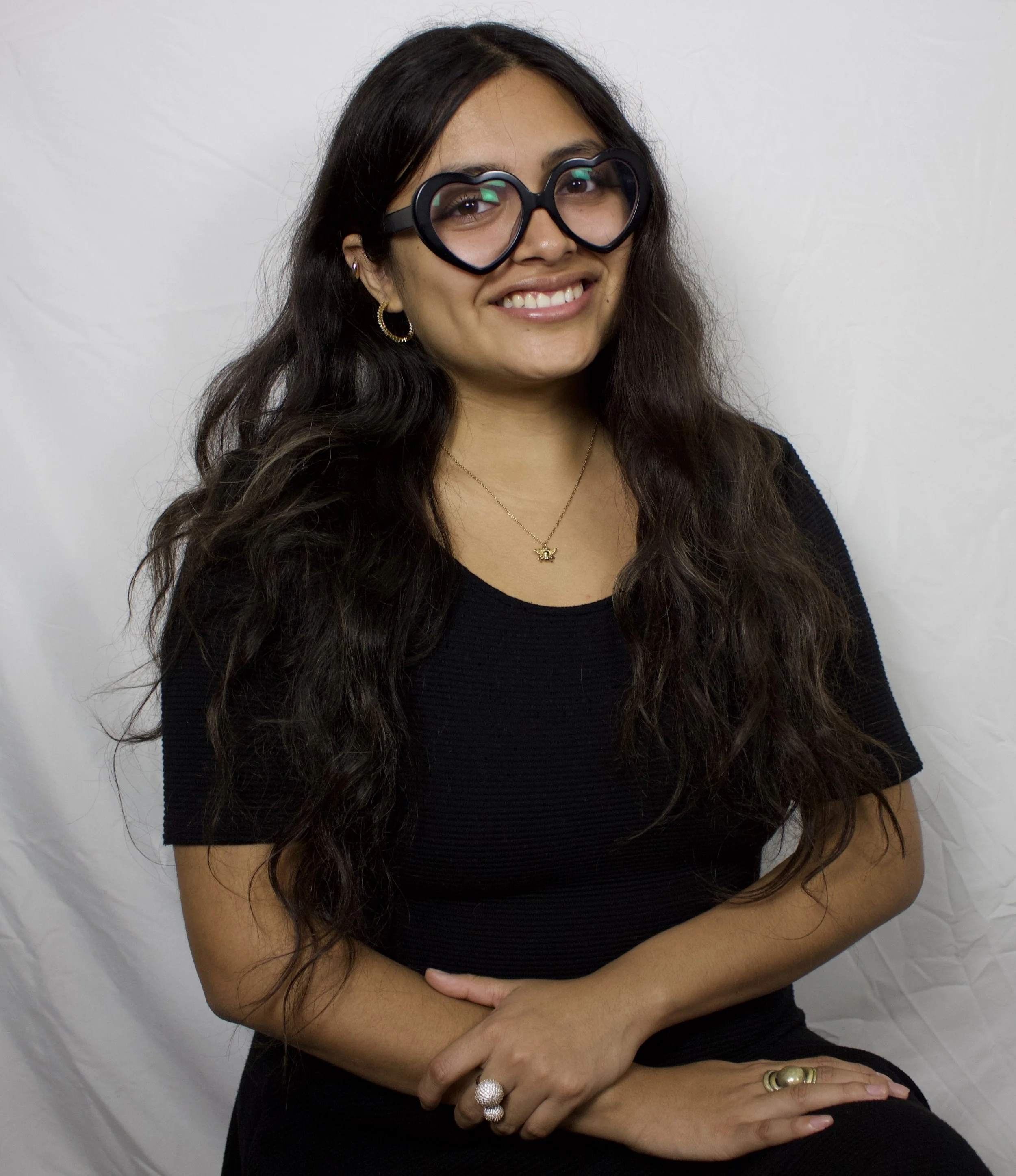 A woman with long dark wavy hair wearing large black heart-shaped glasses, gold hoop earrings, a small bee necklace, and a large pearl ring on her right hand, sitting against a plain white background.