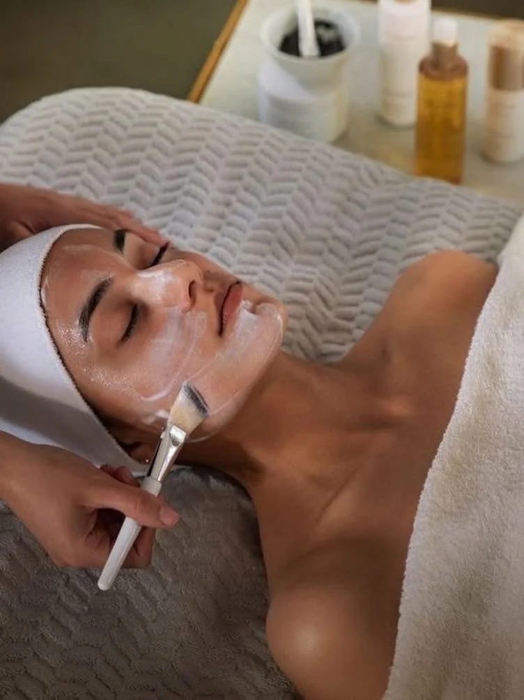 Woman receiving a facial treatment while lying on a bed with a towel covering her body, with skincare products on a table in the background.
