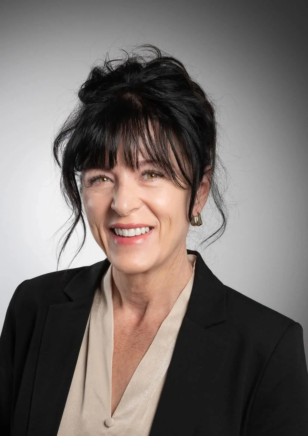 A woman with dark, styled hair and light-colored eyes smiling, dressed in a black blazer and a beige blouse with a collar, against a plain gray background.