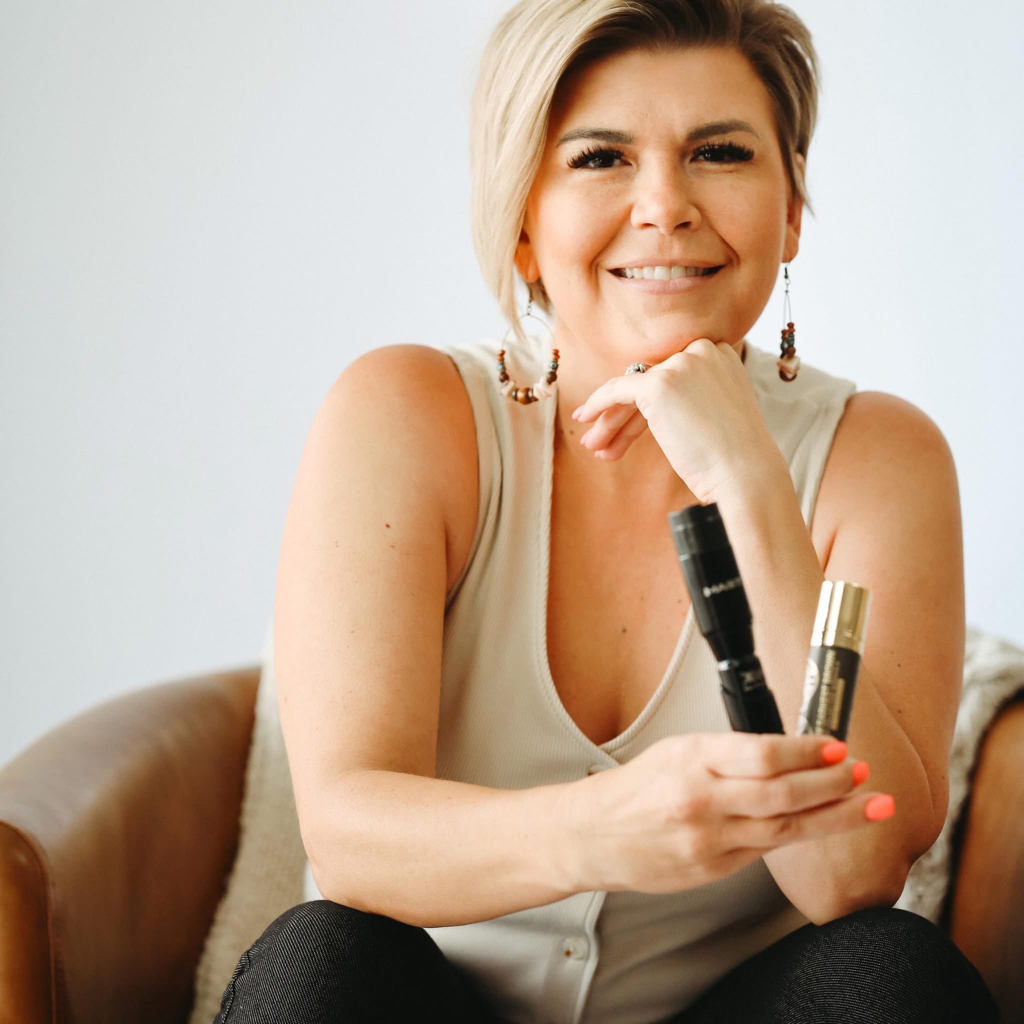A woman with short blonde hair smiling, resting her chin on her hand, wearing earrings, a white top, and makeup, sitting with her elbows on her knees, holding a black marker or pen in her right hand, with a white background.