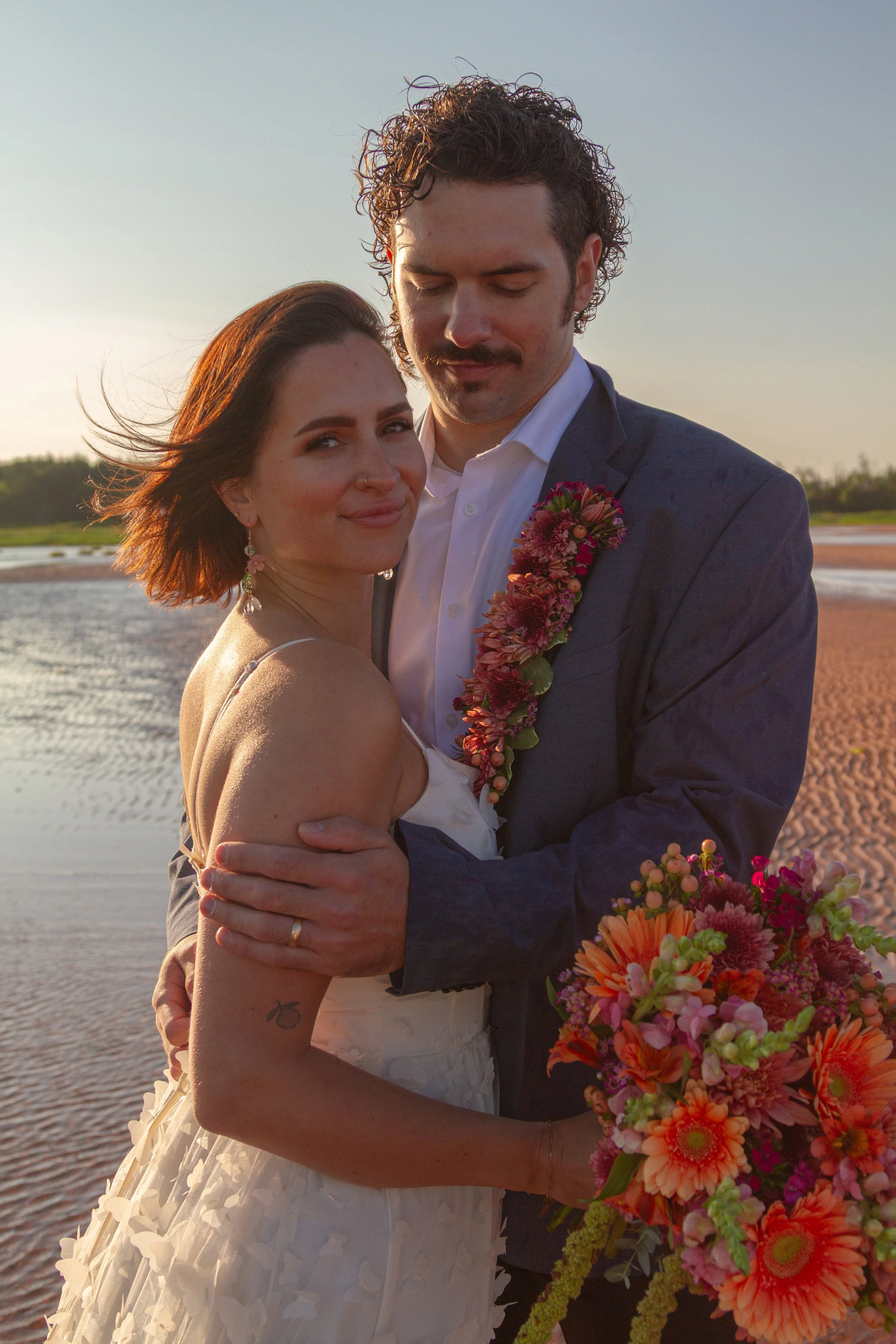 A bride and groom standing close together outdoors at sunset, embracing each other with water and landscape in the background. The bride is smiling gently while the groom has his eyes closed and is holding a colorful bouquet of flowers.