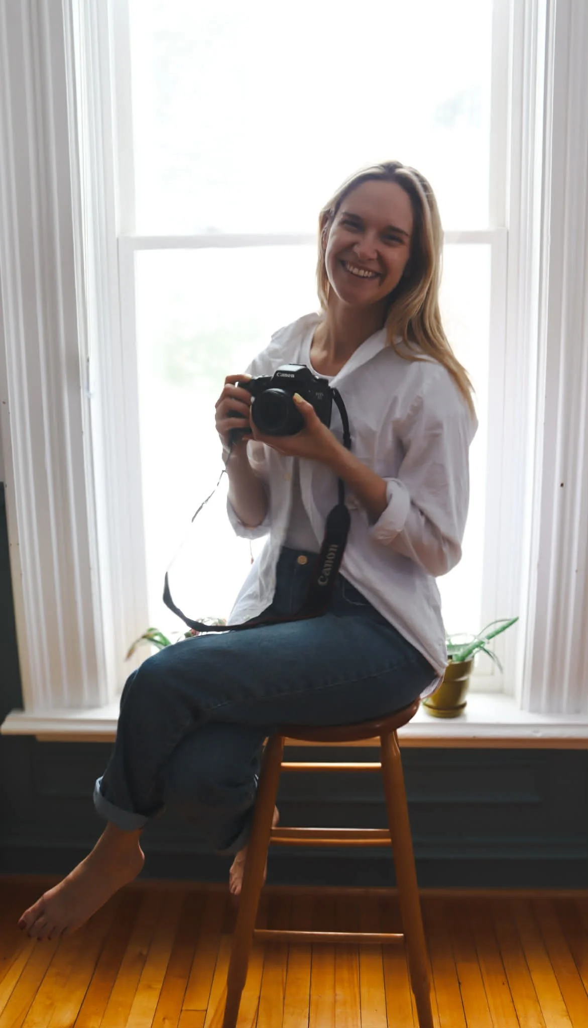 A woman with long blonde hair smiling while sitting on a wooden stool in front of a large window, holding a Canon camera, wearing a white shirt and dark jeans, with a houseplant in a yellow pot nearby.