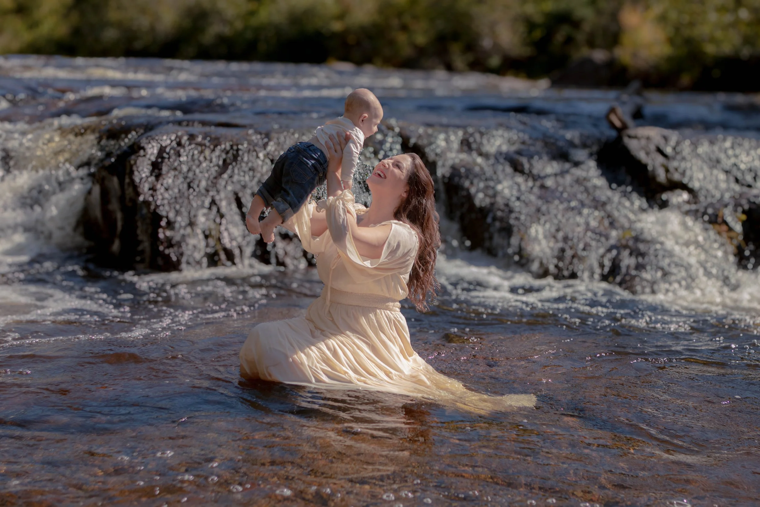 A woman in a cream-colored dress is kneeling in a river, holding a smiling baby dressed in a light shirt and dark jeans above her head, with water splashing around them.