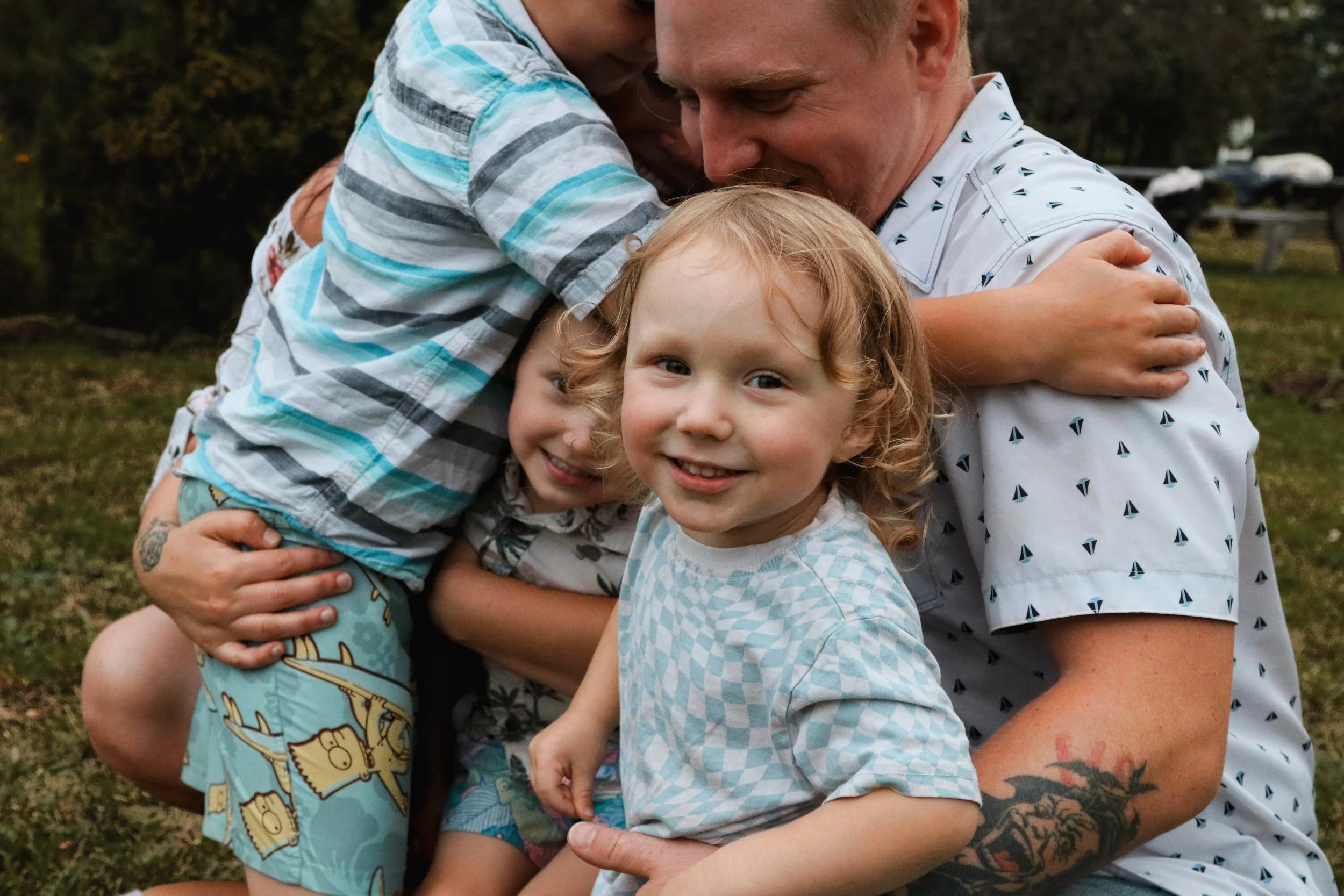 A man with a tattoo on his arm hugging two young children, one with red hair and the other with short blonde hair, outdoors with trees and grass in the background.