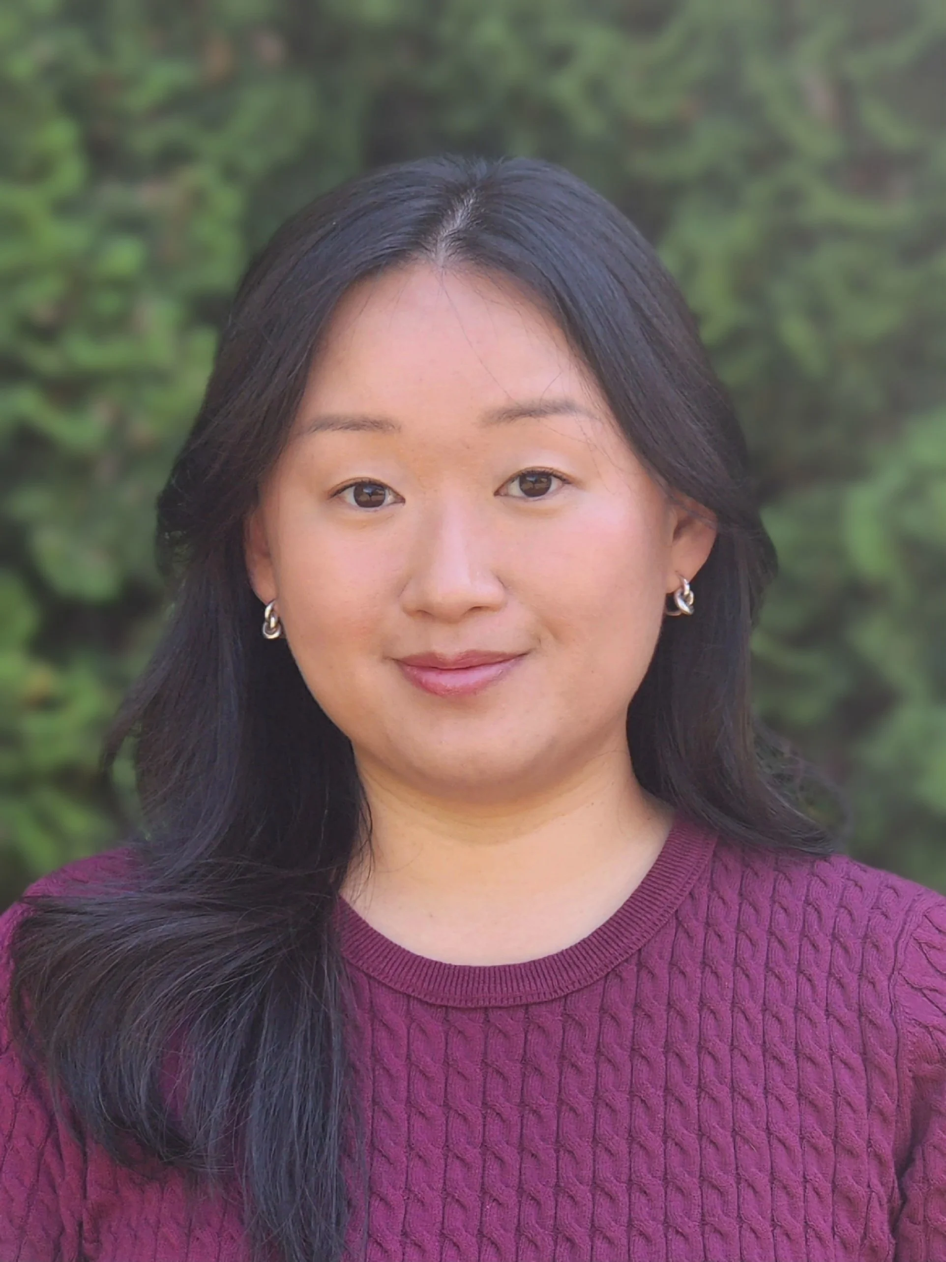 Close-up of a smiling woman with long dark hair wearing a maroon sweater and silver hoop earrings, standing outdoors with green foliage in the background.