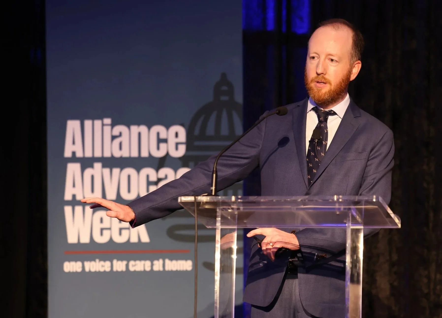 A man with a beard and a suit speaking at a podium during a conference with a large screen behind him with a logo of a dome building.