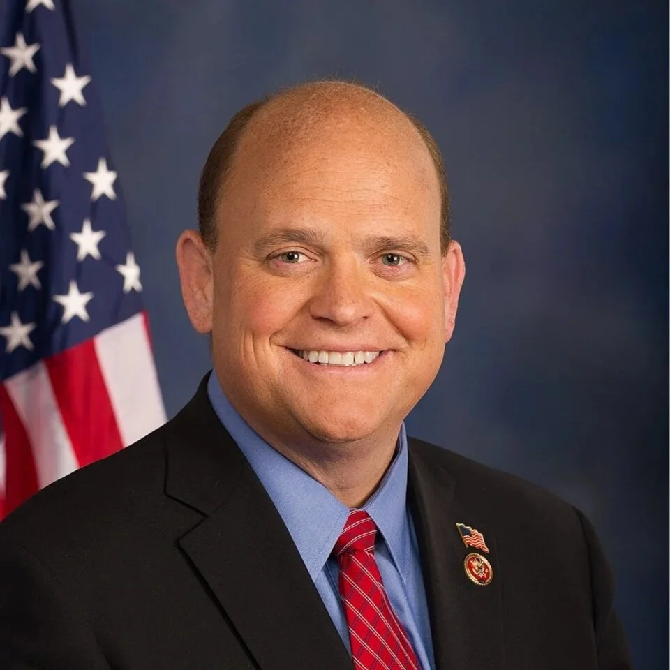 A man in a suit and red tie smiling in front of an American flag.