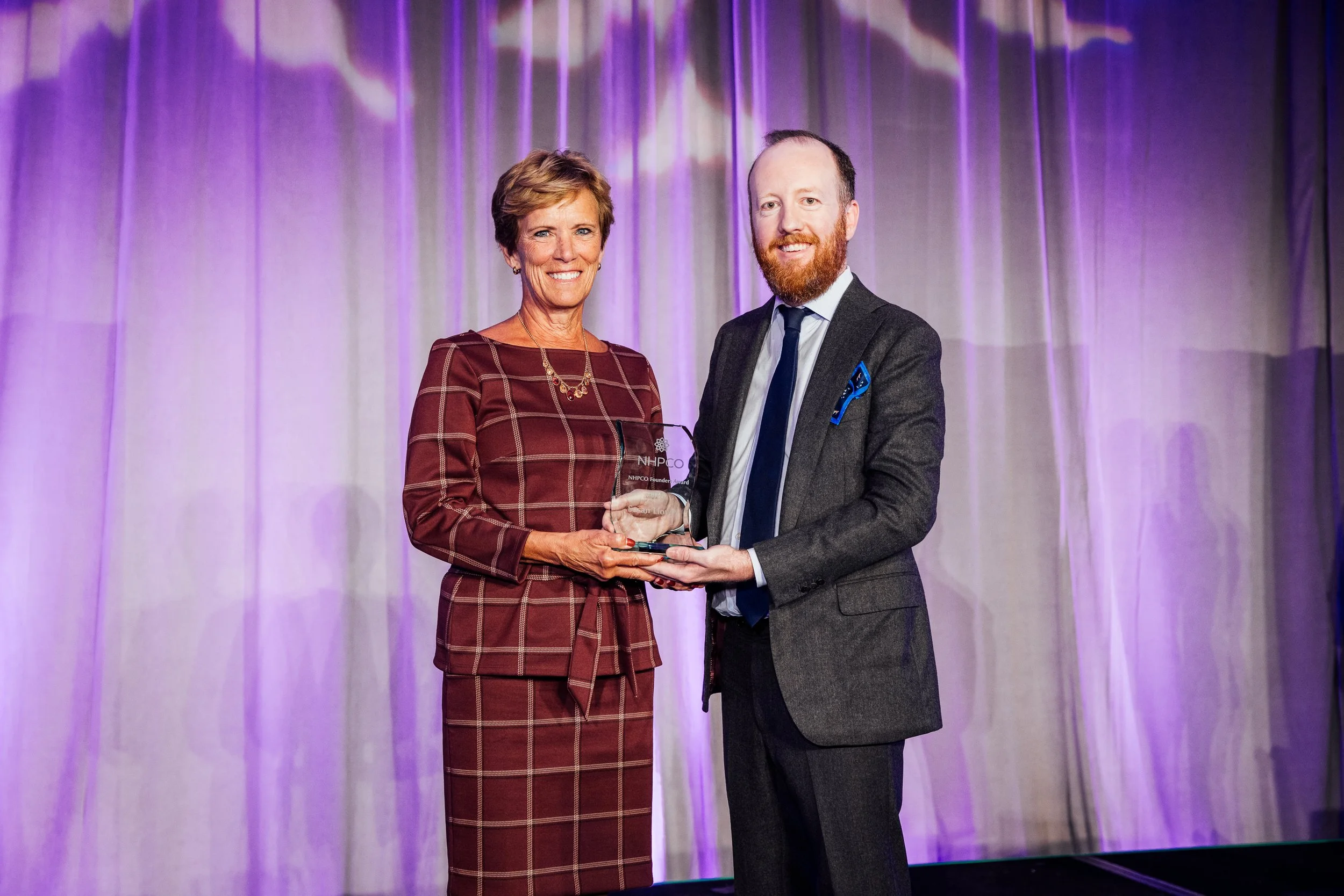 A woman with short light brown hair wearing a burgundy plaid dress is receiving an award from a man with a beard, wearing a dark gray suit and blue tie, on stage with purple curtains in the background.