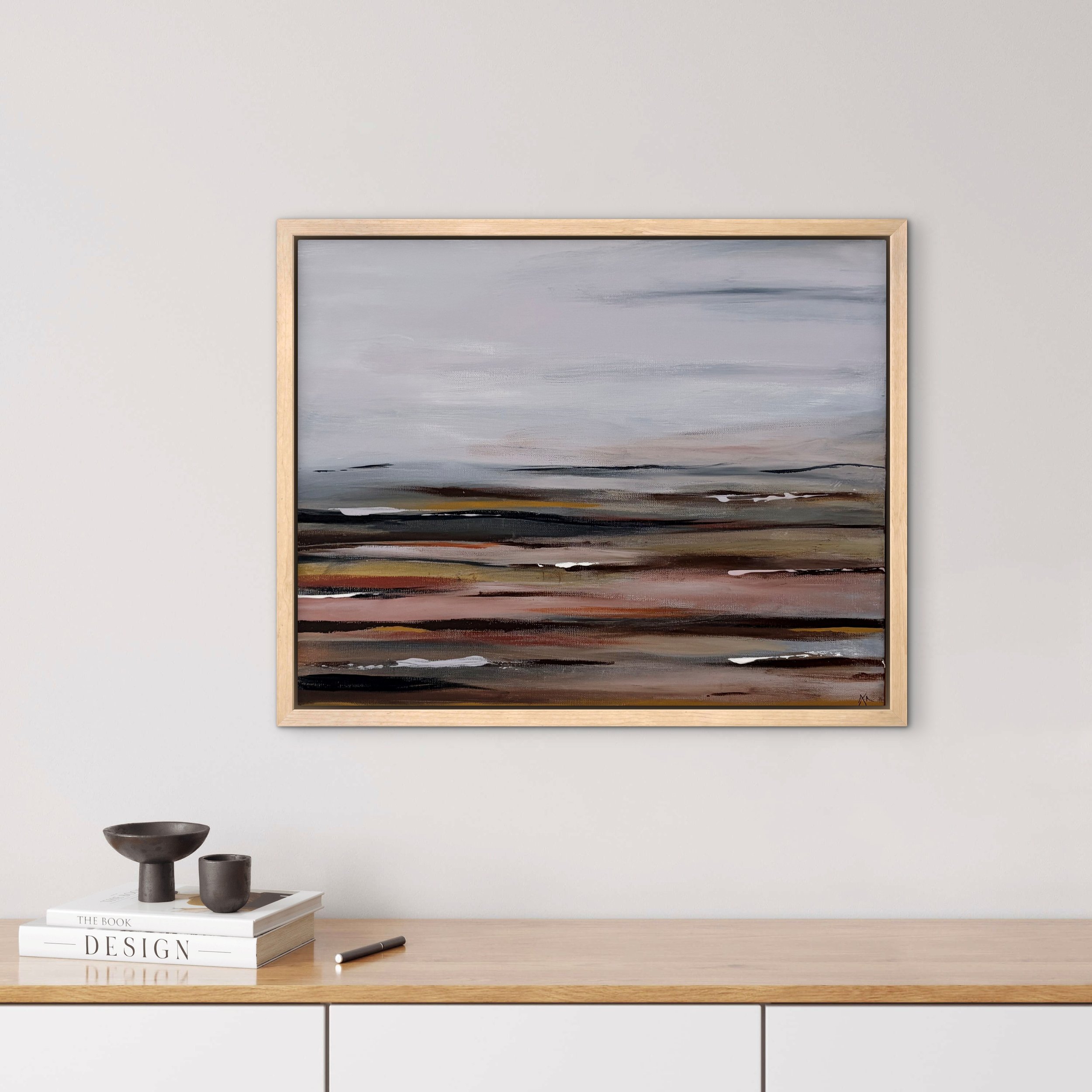 A minimalist room interior with a light-colored wall, a large framed abstract landscape painting, a white table holding a black ceramic bowl, a smaller cup, a book titled 'The Book of Design,' and a black pen.