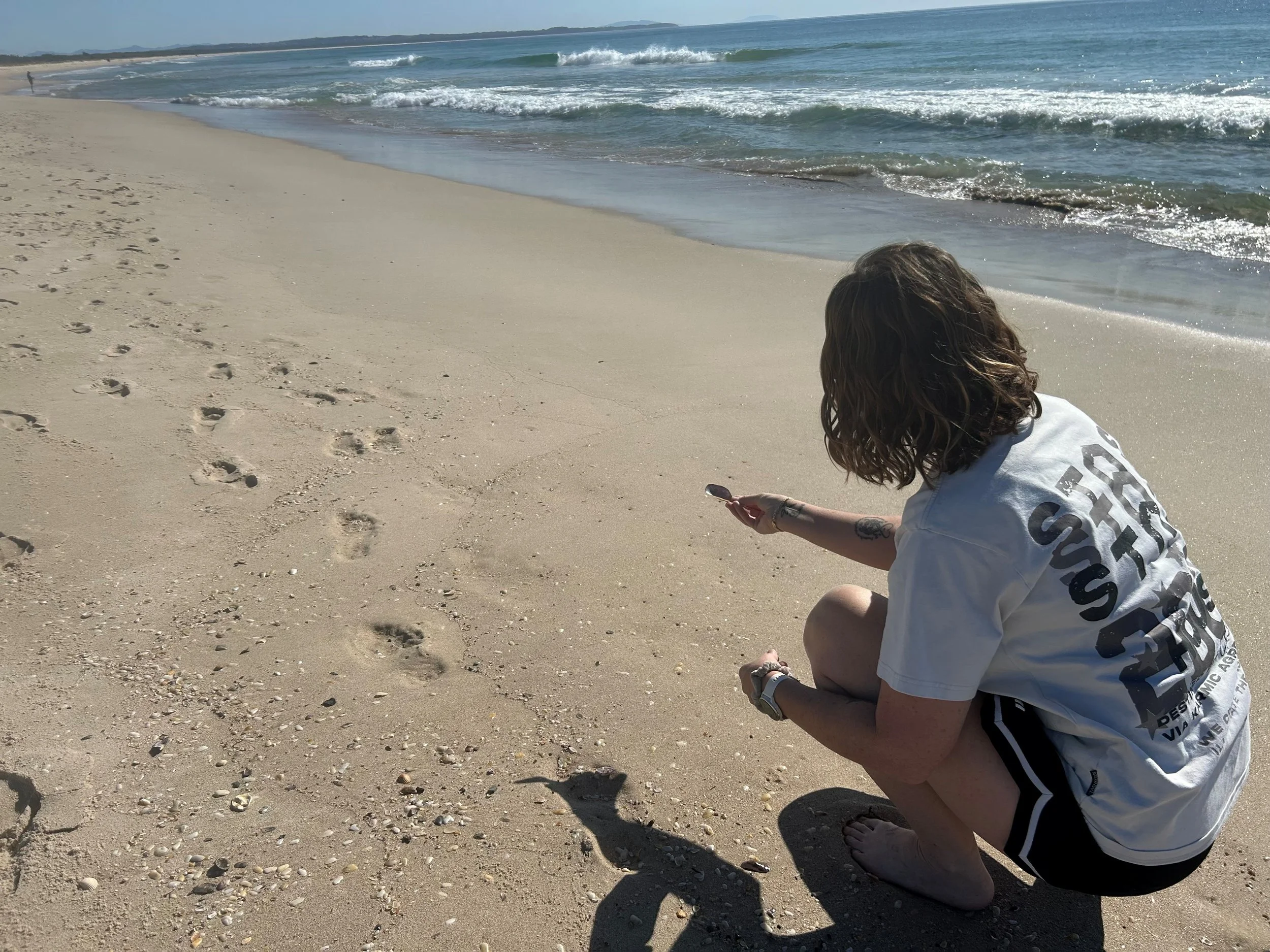 Person with wavy hair crouching on sandy beach looking at seashells, shoreline with gentle waves and distant land visible in background.