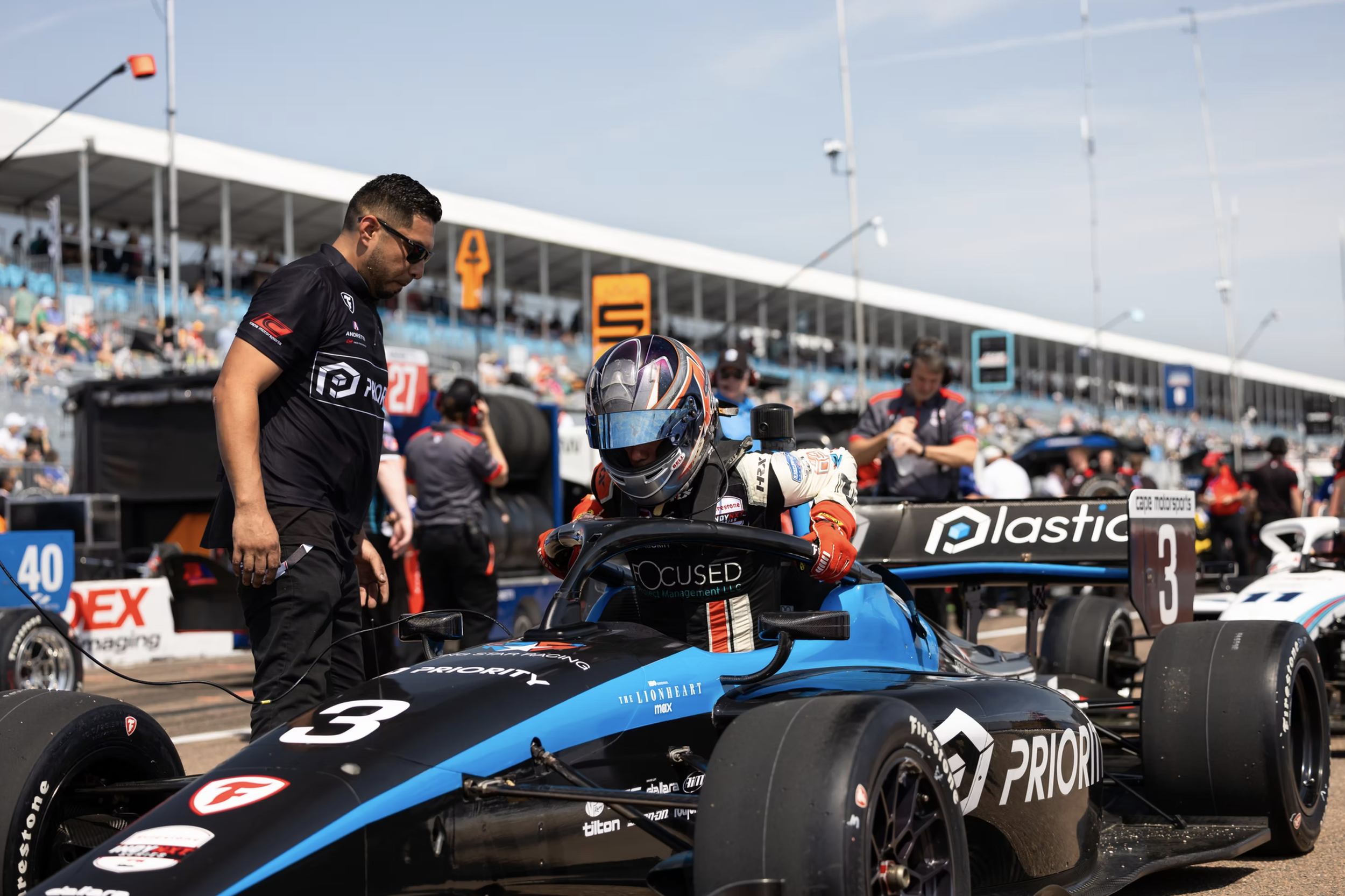 Michael d'Orlando driving professional in a helmet and racing suit sitting in a black and blue open-wheel race car with team staff around on a race track. Spectators in the background.