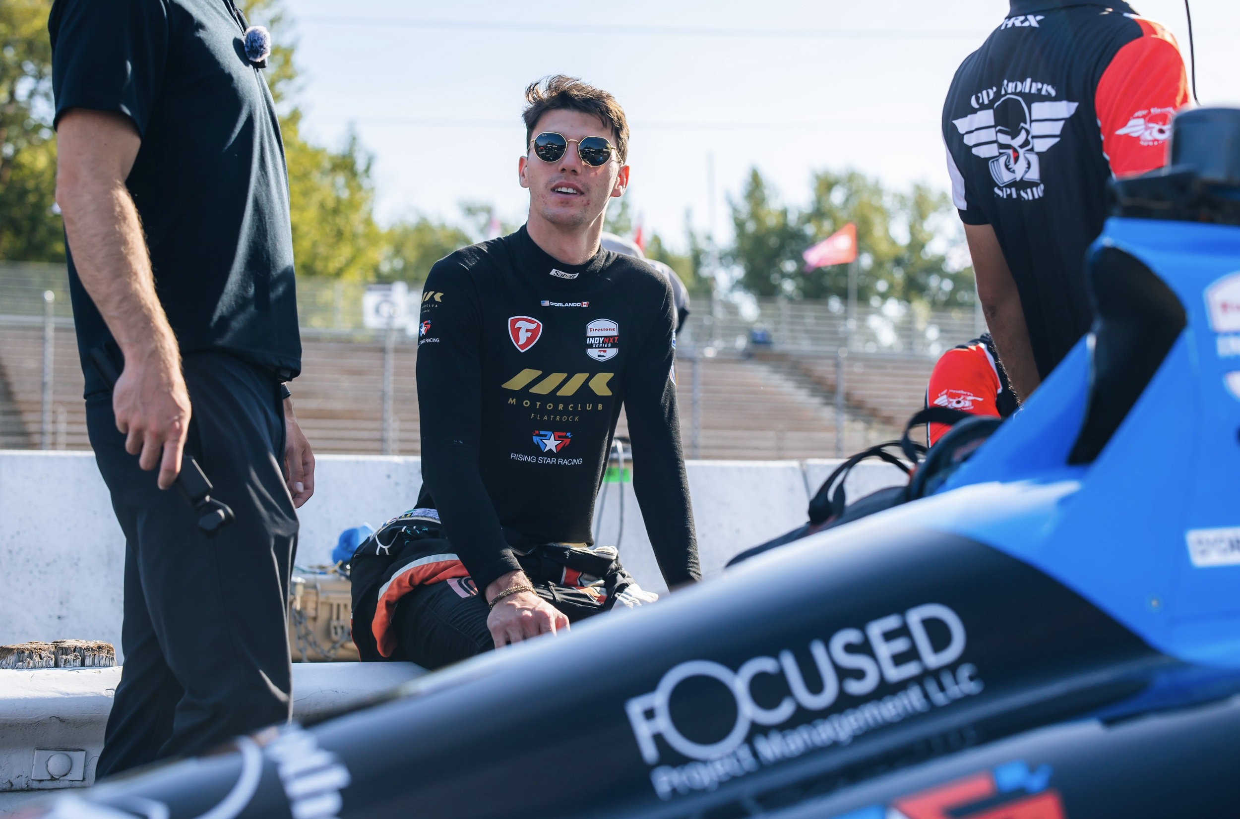 A man wearing sunglasses and a racing suit sitting on a bench at a race track, with two men standing next to him. The scene is outdoors with some trees and flags in the background.