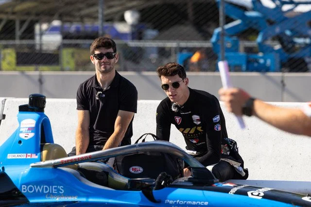 Two men in racing suits and sunglasses sitting next to a blue IndyCar racing vehicle on a racetrack.