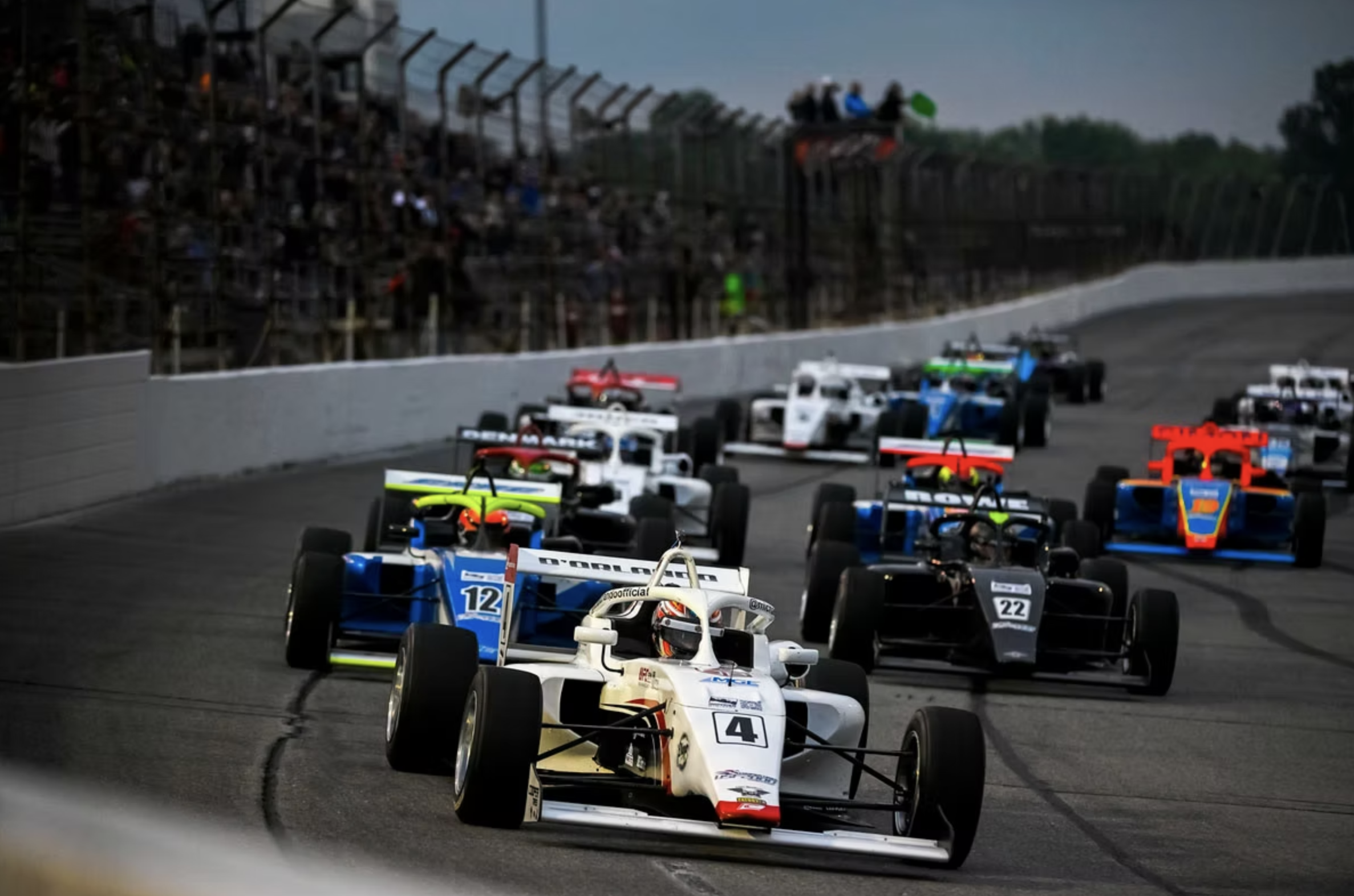 Michael d'Orlando and multiple race cars in a motorsport race on a track with a dark cloudy sky, spectators watching from grandstands in the background.