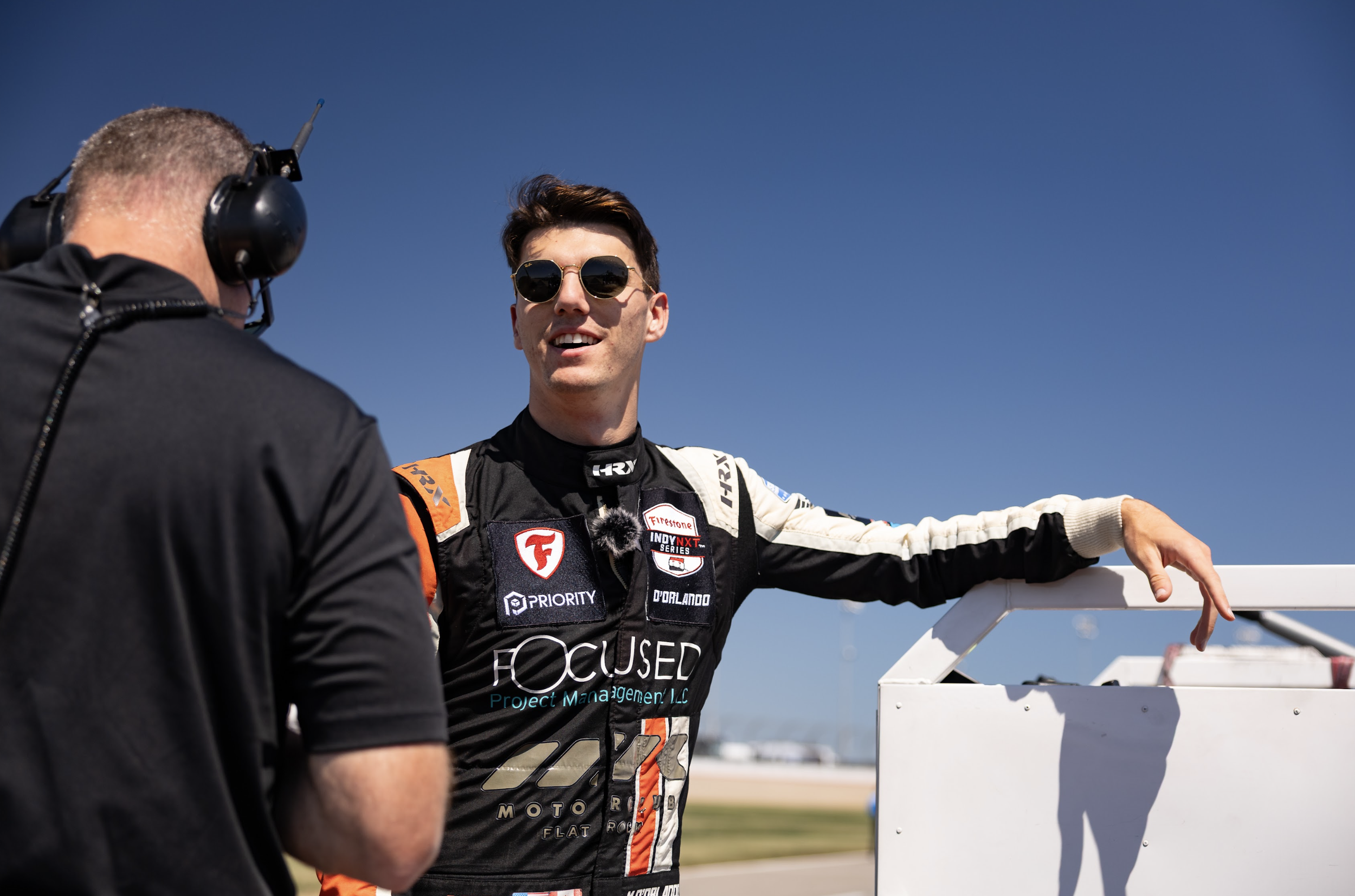 A race car driver wearing sunglasses and a racing suit with various sponsor logos, standing by a white fence on a race track while talking to another person wearing a black jacket and headphones, under a clear blue sky.