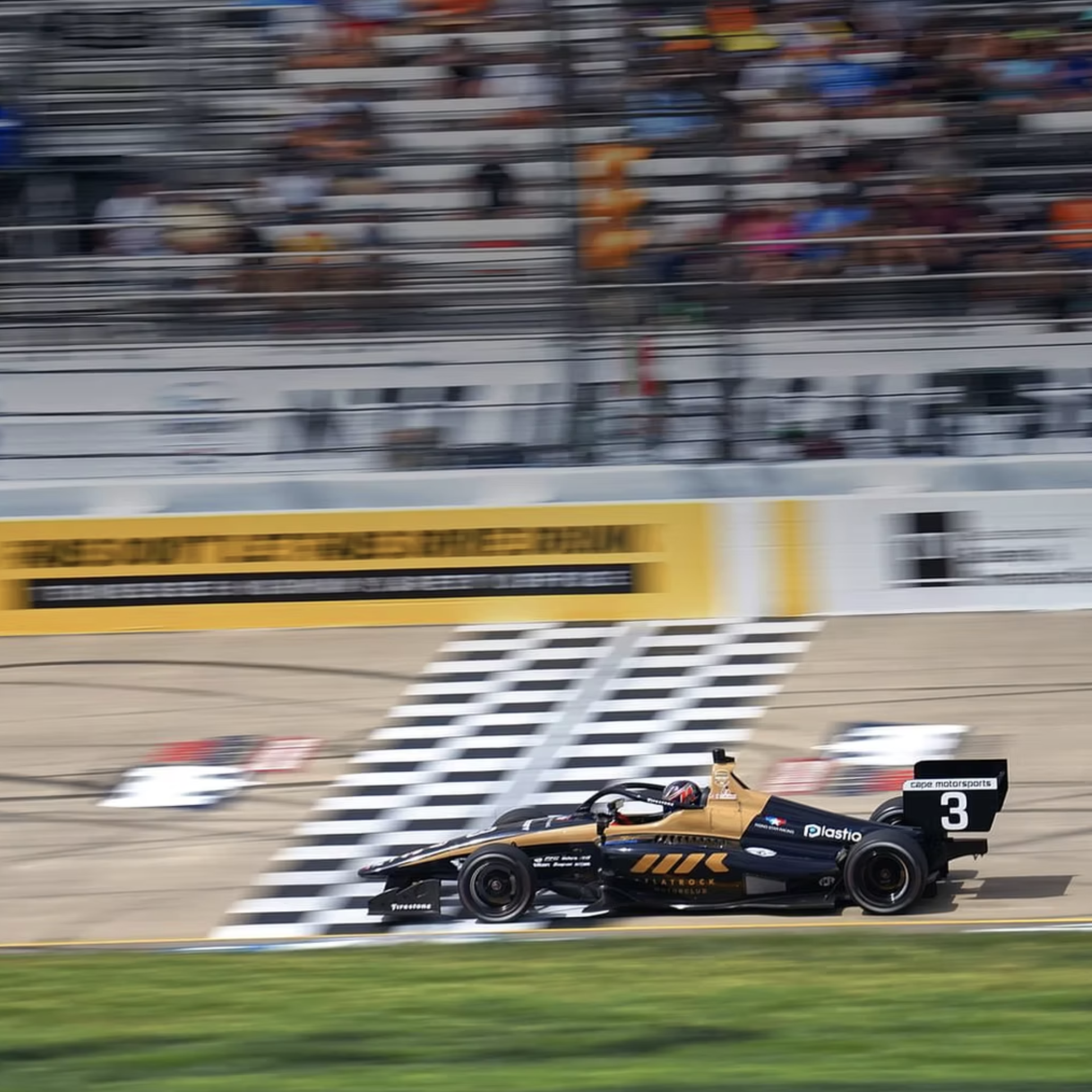 Michael d'Orlando blurred image of a race car on a race track, with a focus on a black and gold race car in the foreground and a crowd of spectators in the background.
